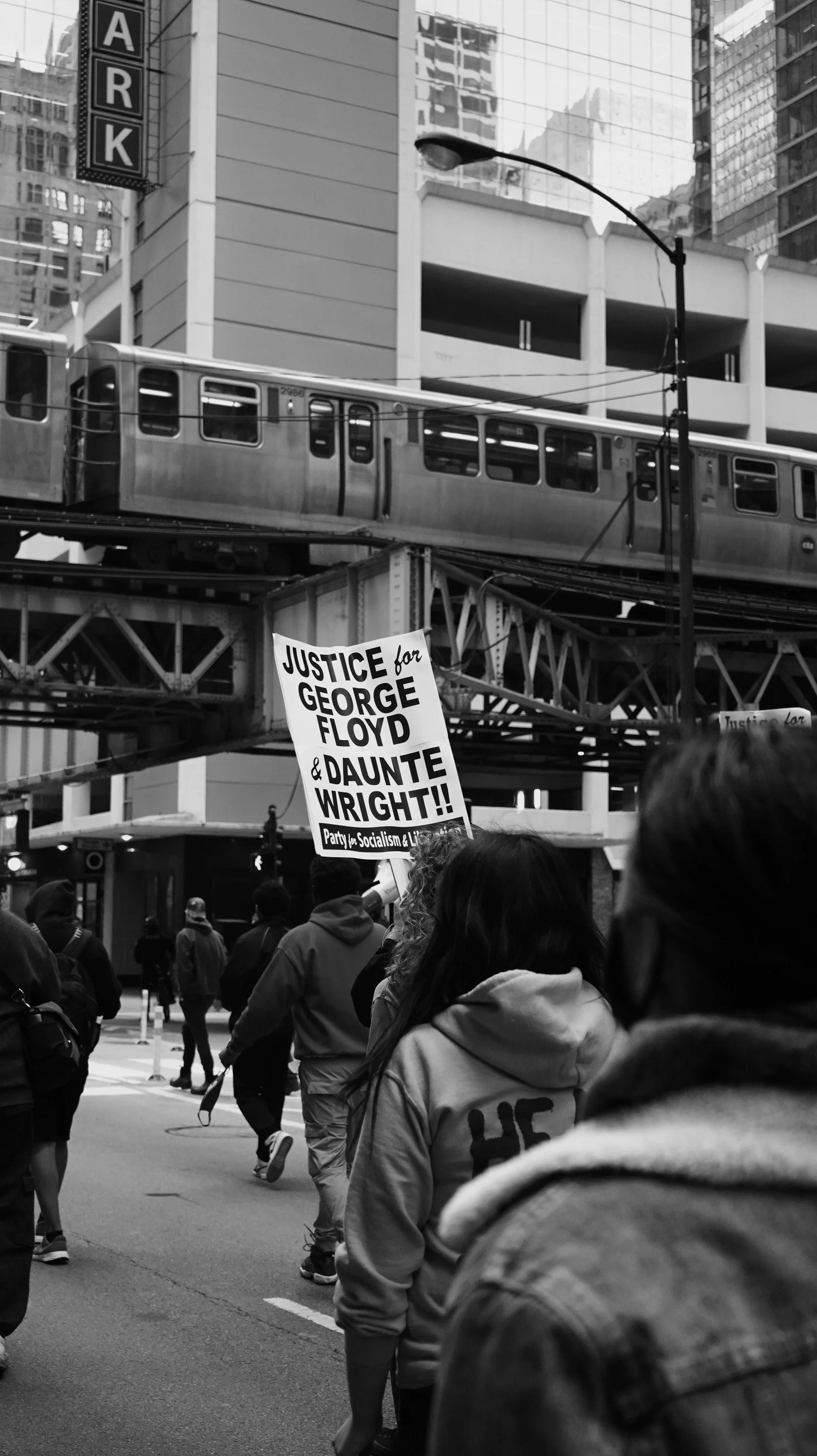 A black and white photo of a protest march in an urban area with tall buildings and a train above the street. A sign held by one of the protesters reads, 'Justice for George Floyd & Daunte Wright!! Party for Socialism & Liberation.'