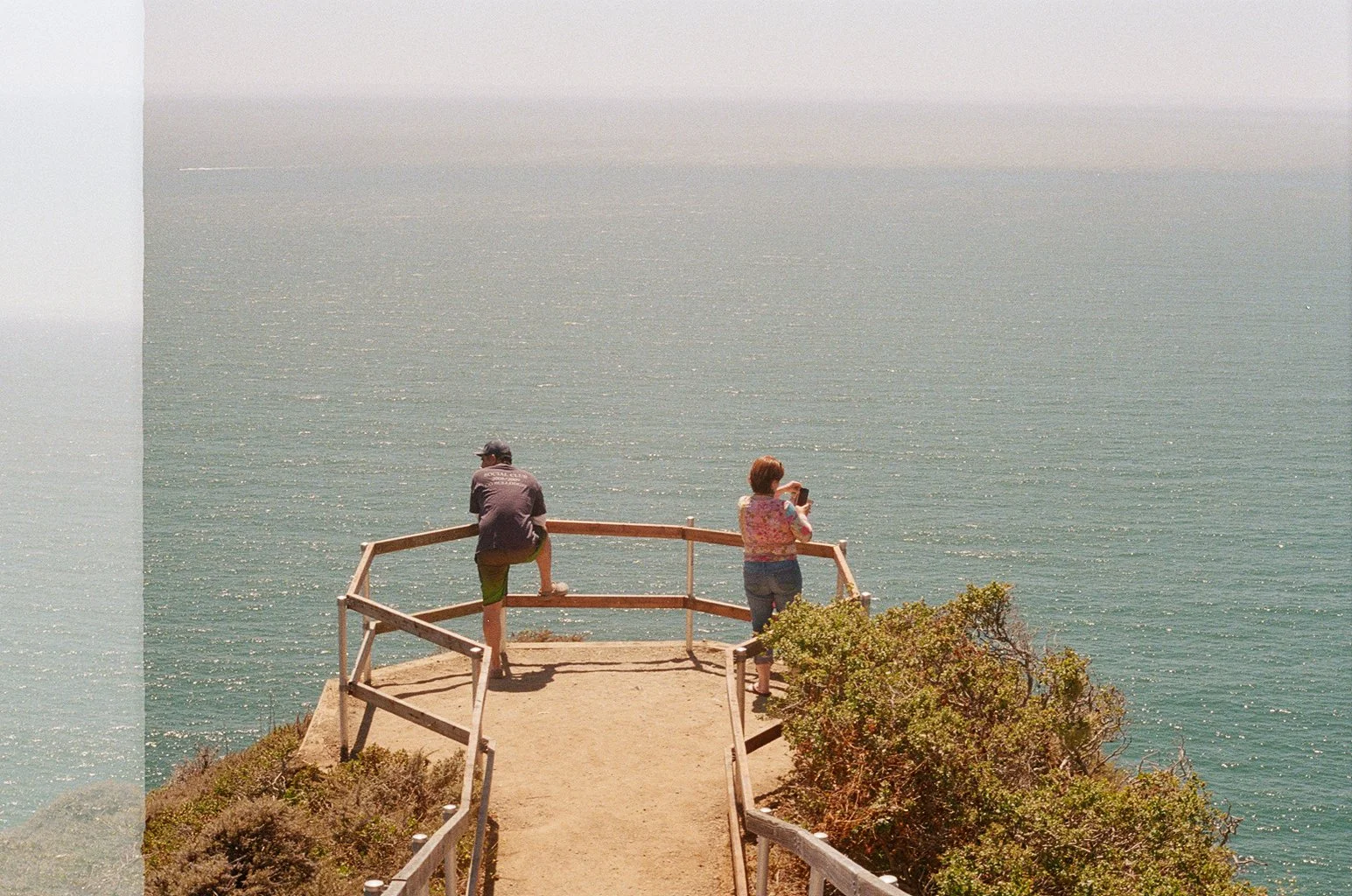 A man and a woman are standing on a wooden observation deck overlooking the ocean, with one woman taking a photo and a man looking out at the water.