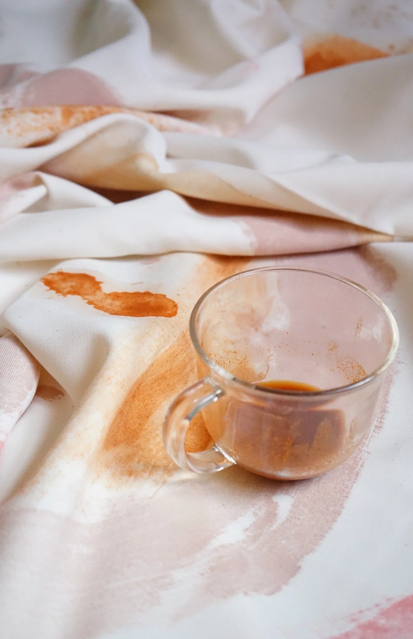 A small glass cup with remnants of coffee, placed on a crumpled white and pink cloth with watercolor-like stains.