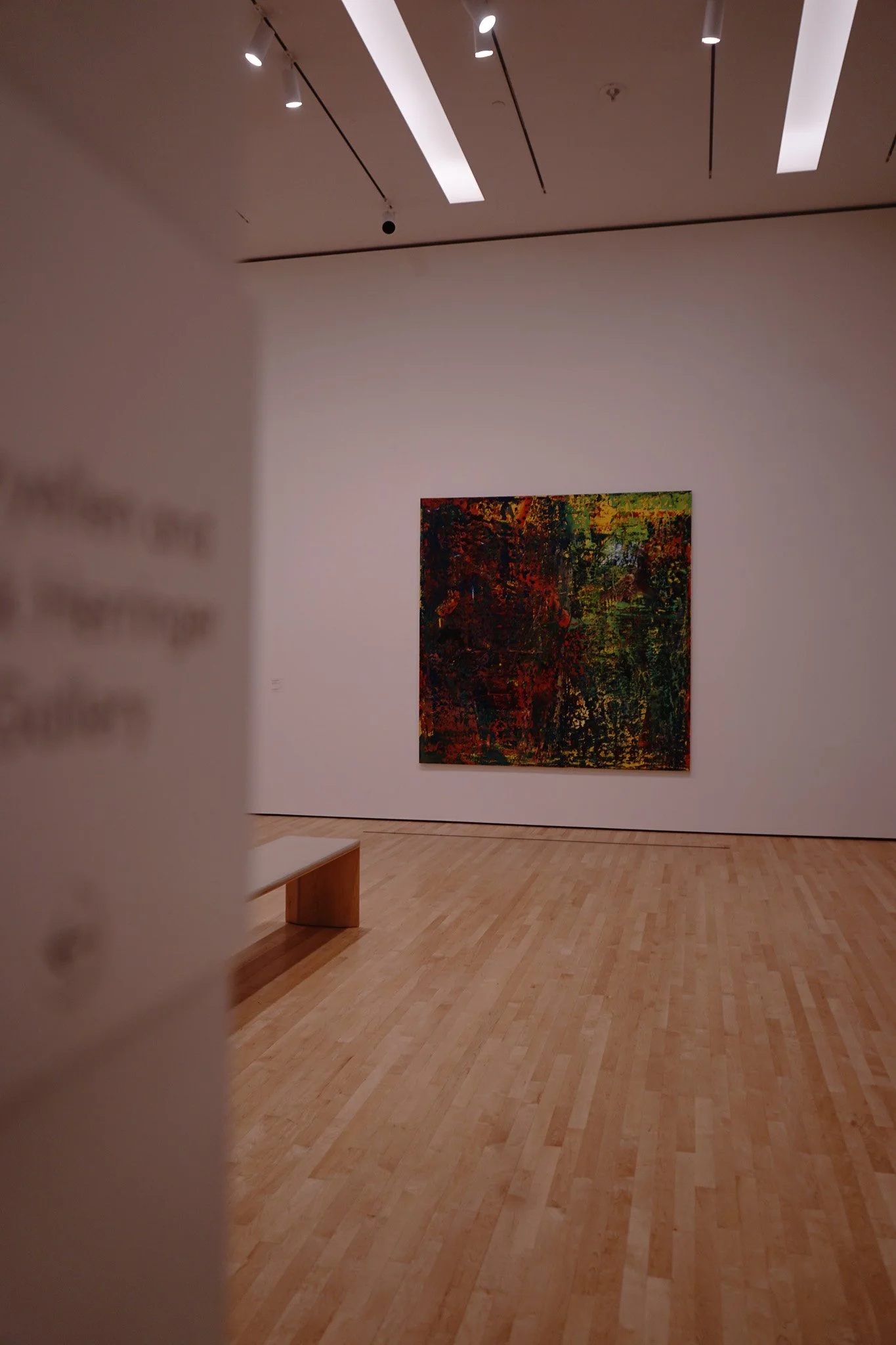 An art gallery with a colorful abstract painting on a white wall, a wooden bench, and a wooden floor.