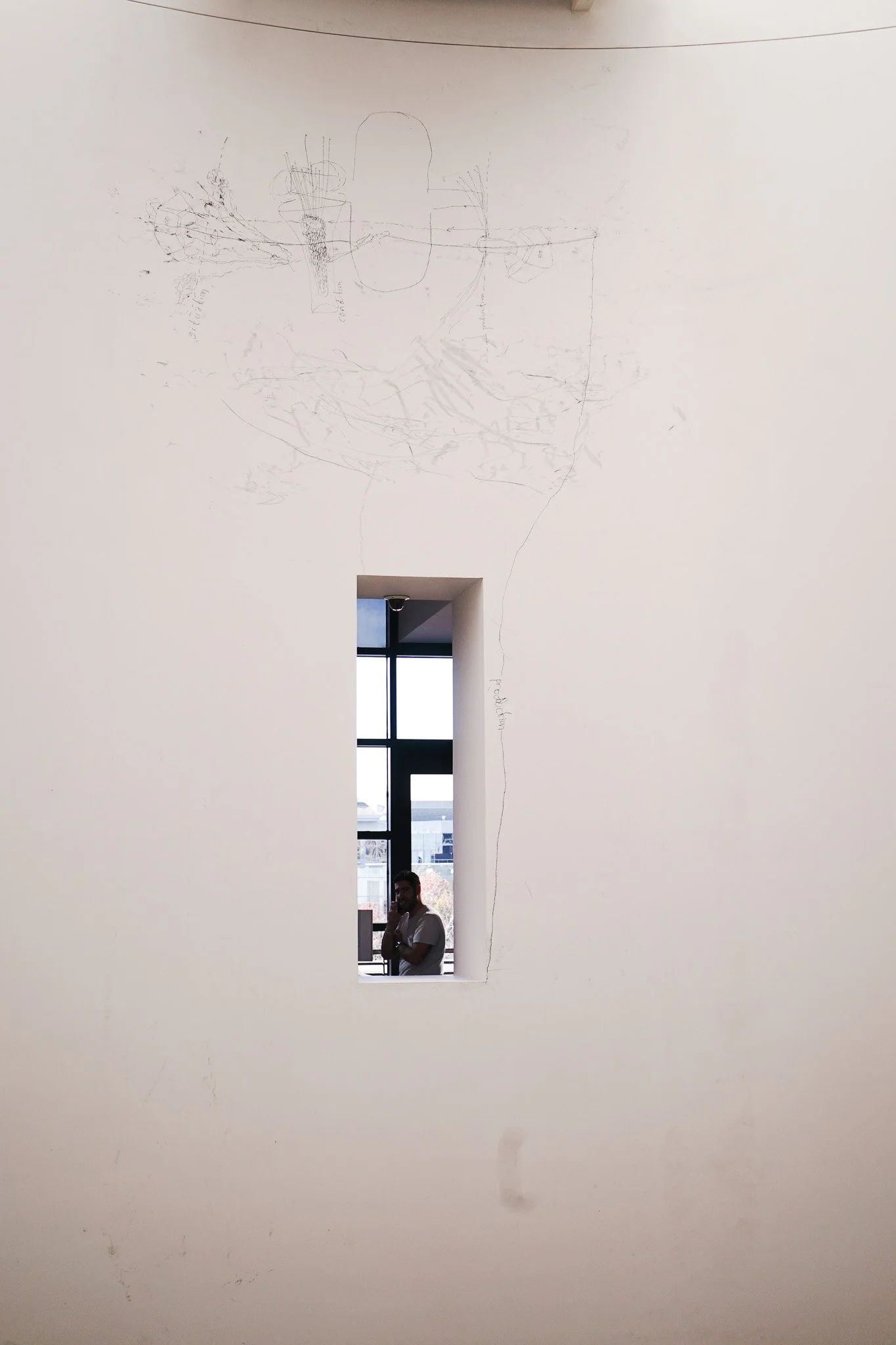 White wall with a rectangular cutout window, through which a person is visible standing in front of a large window, and faint sketches are made on the upper part of the wall.