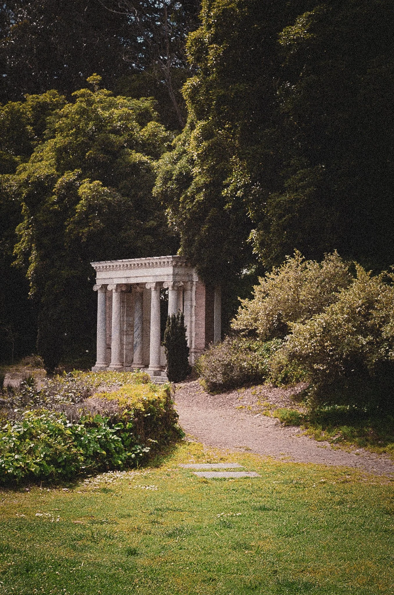 A garden with a small classical temple structure surrounded by lush greenery and trees, with a dirt pathway leading to it.