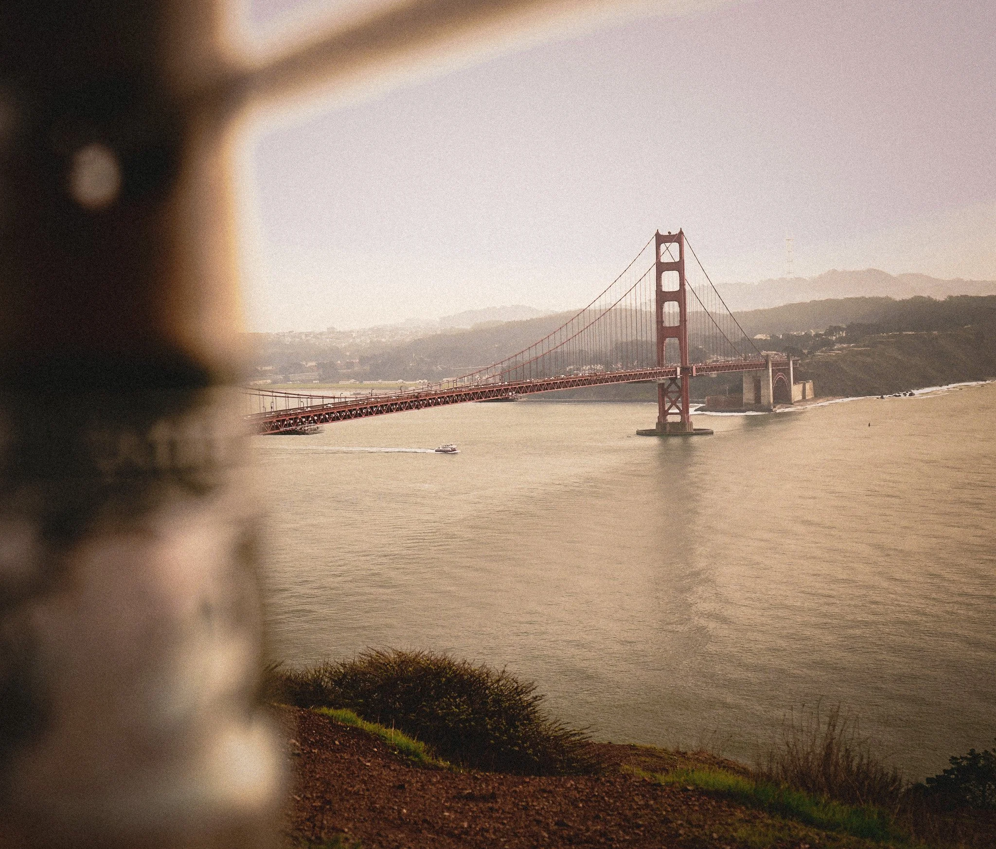 The Golden Gate Bridge over water with hills in the background, partially obscured by a blurred foreground element.