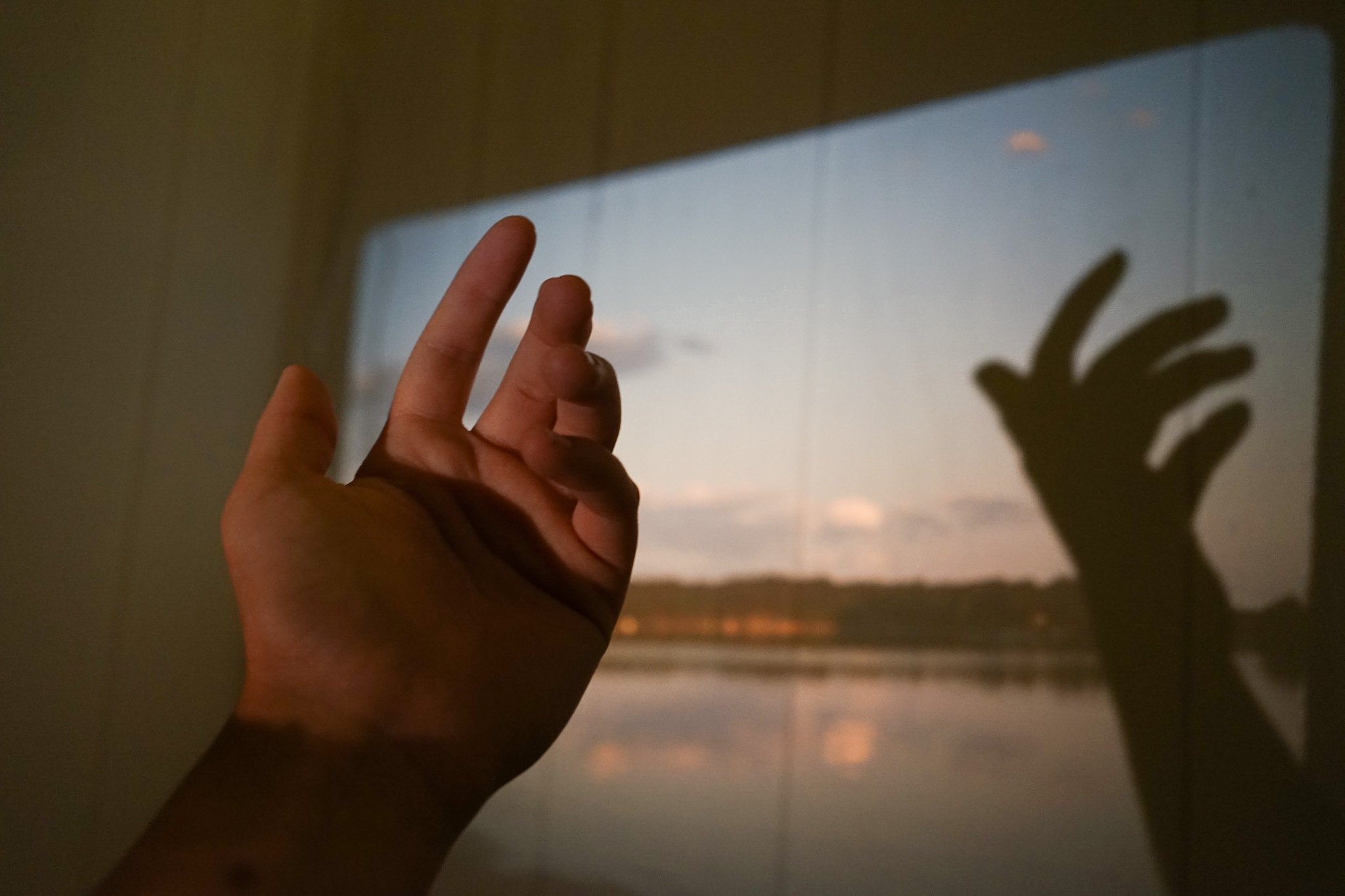A person's hand making an OK gesture with the thumb and index finger touching, casting a shadow on the wall, with a sunset view through a window in the background.