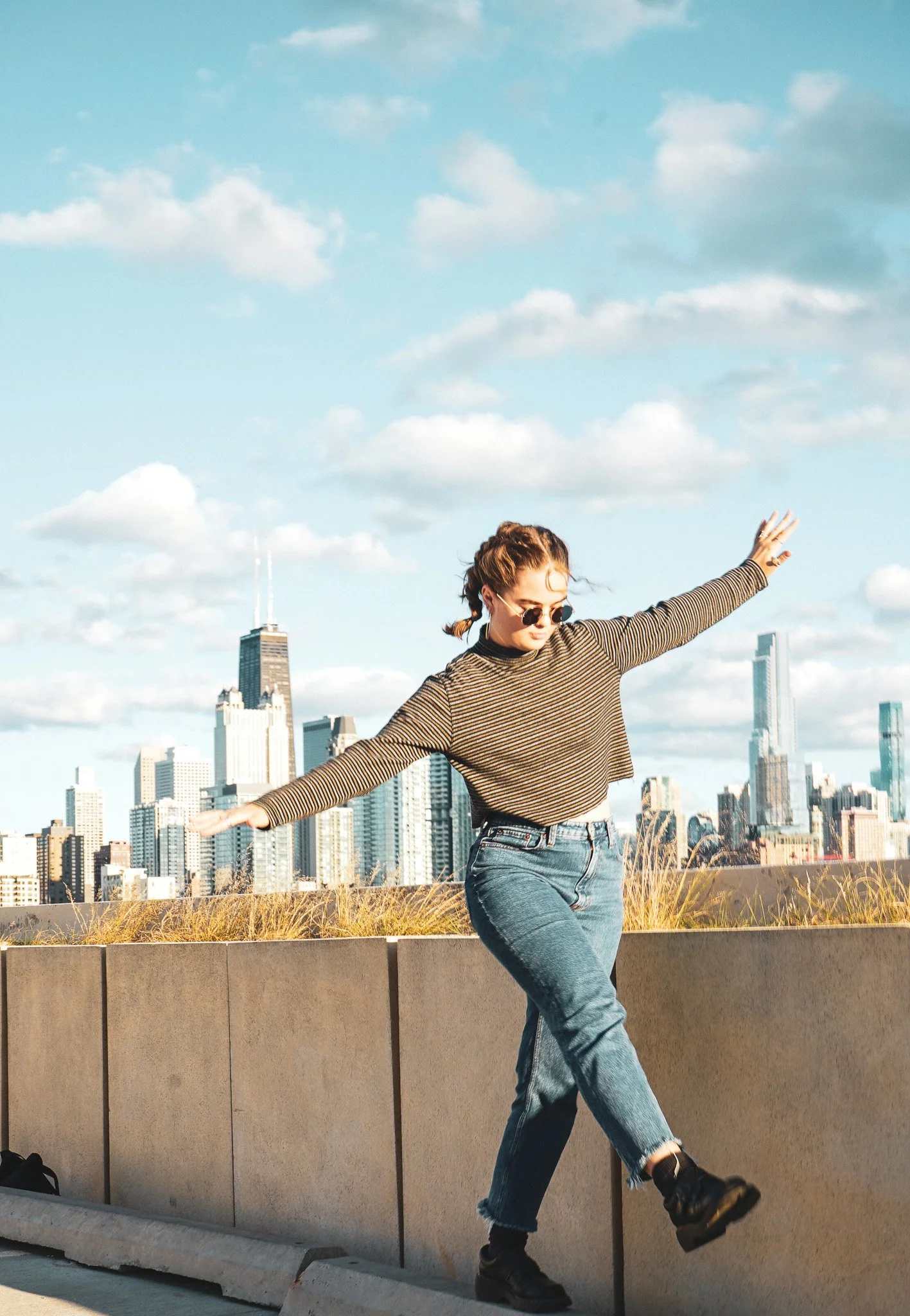 A woman is balancing on a low wall with her arms extended, wearing sunglasses, a striped long-sleeve shirt, jeans, and black boots, with a city skyline in the background under a partly cloudy sky.