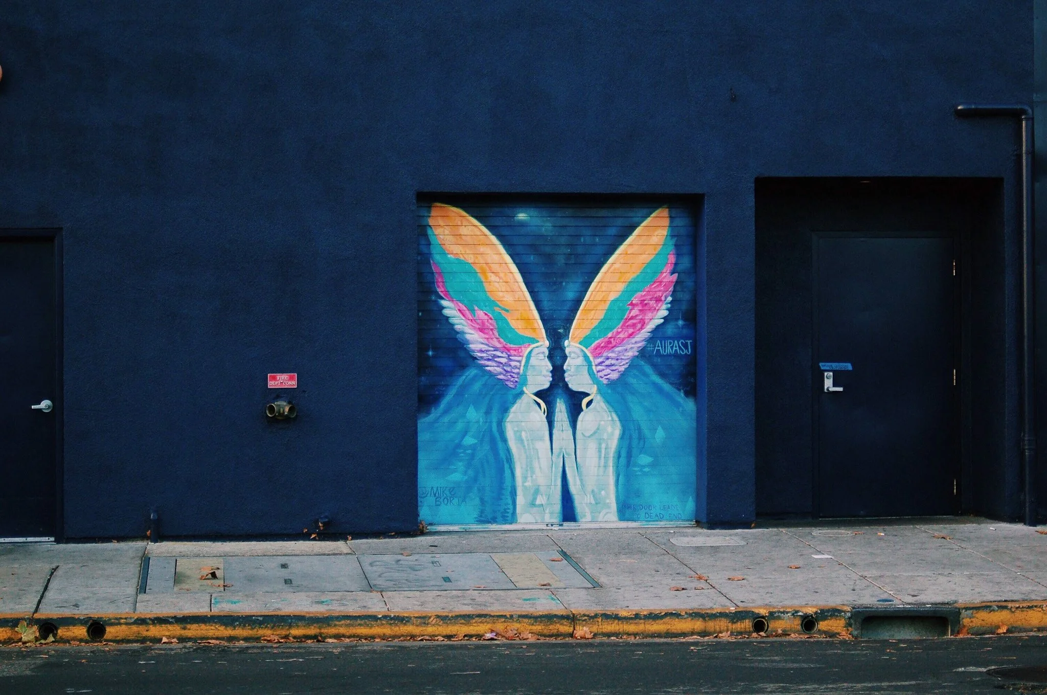 Colorful mural of a symmetrical pair of winged figures facing each other on a dark blue building wall.