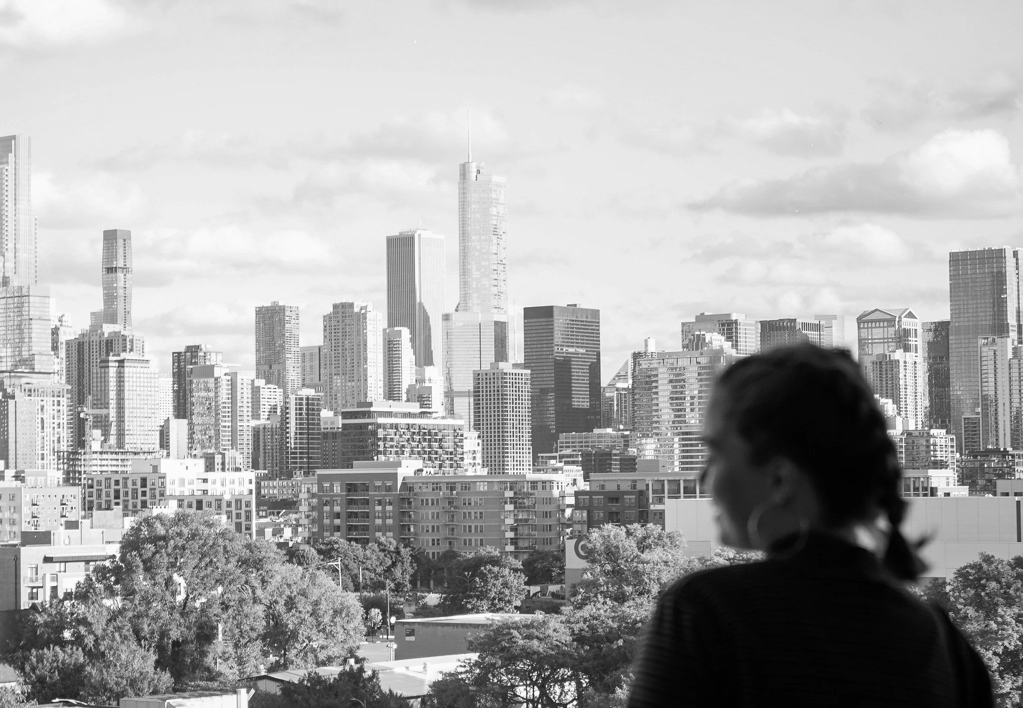 A woman with glasses and curly hair wearing a striped shirt, standing outdoors with the city skyline in the background, including tall skyscrapers and trees.