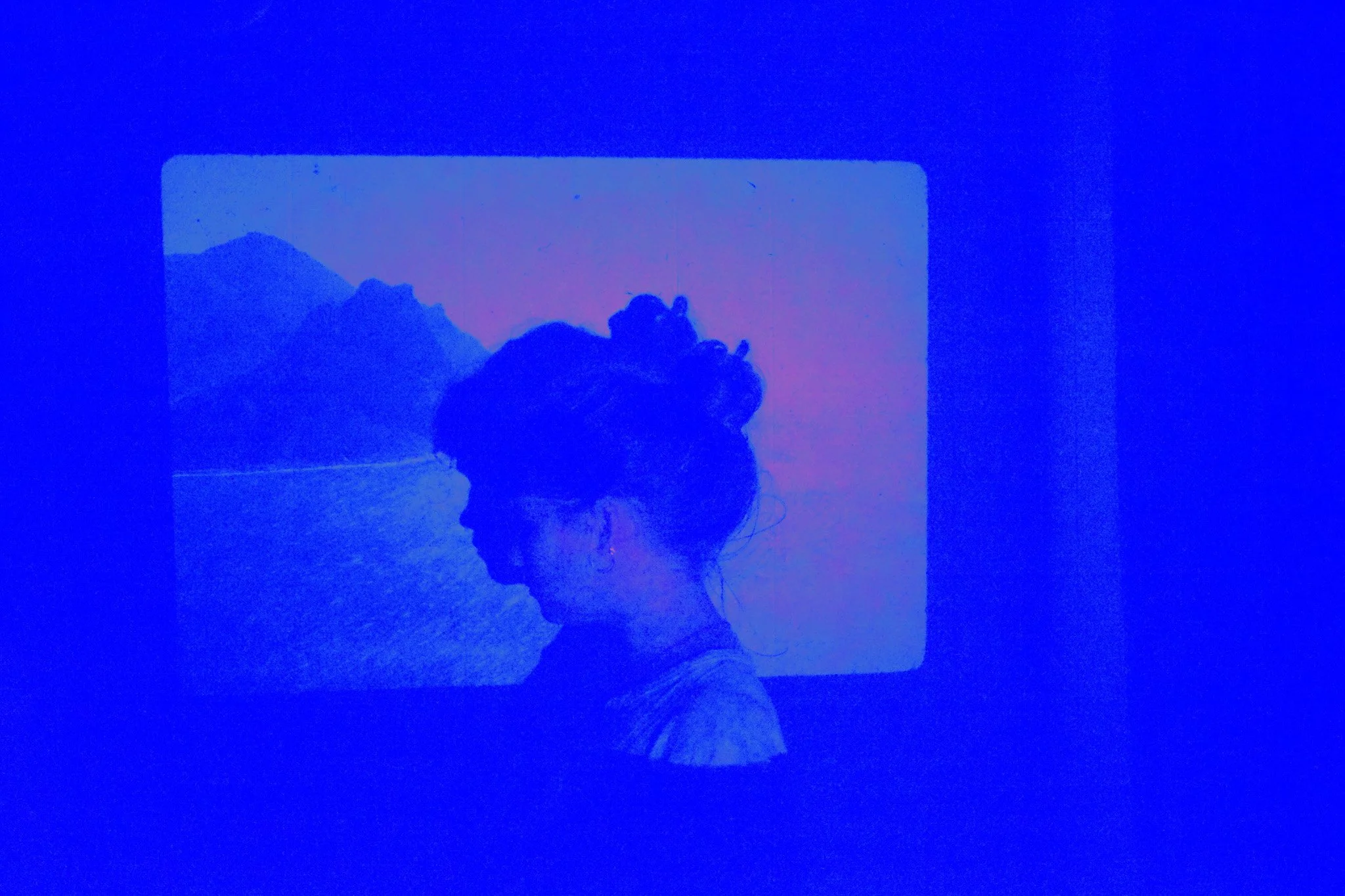 A woman with her hair in a bun is looking downward, inside a framed display showing a scenic mountain landscape, with a pink and purple sky.