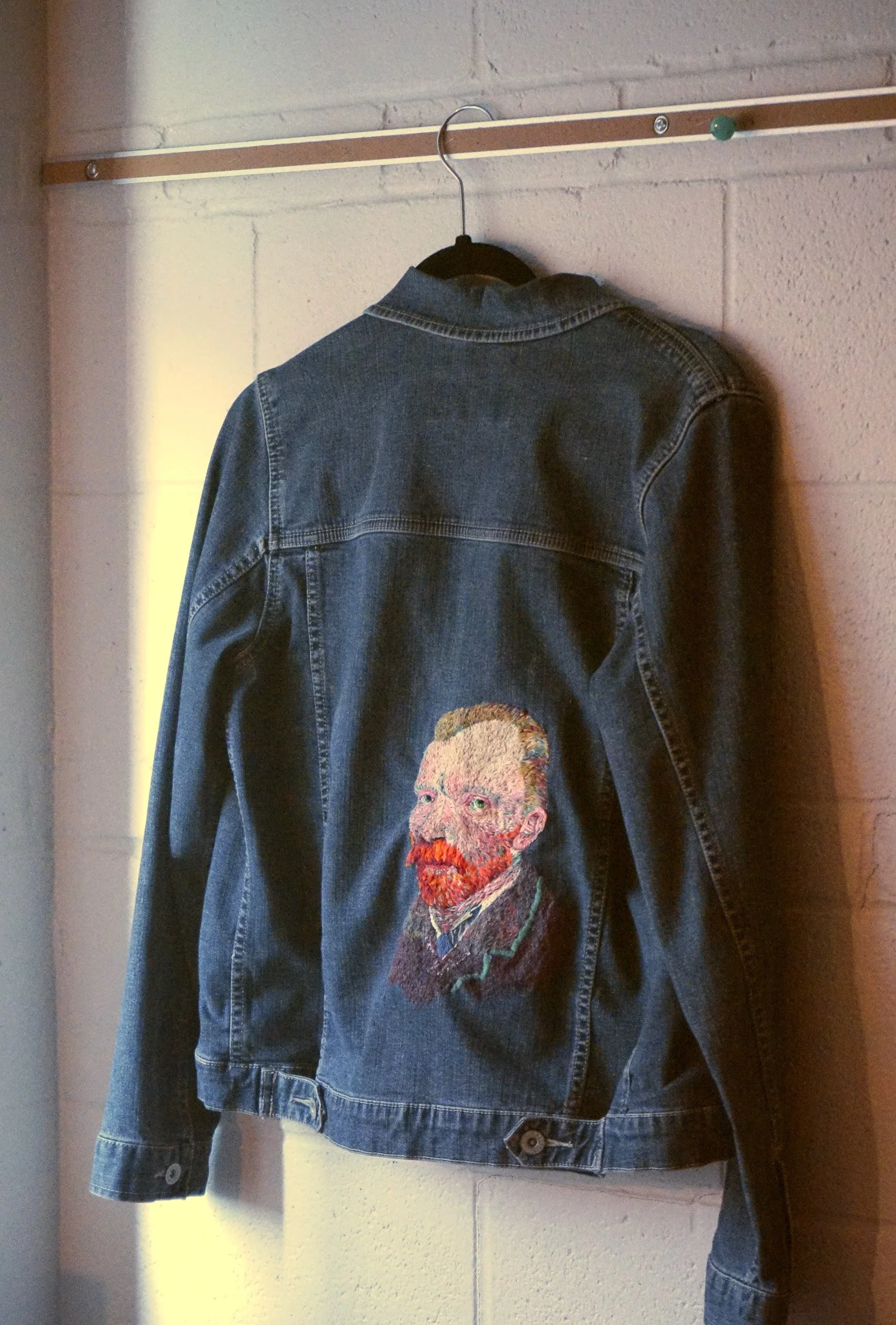 Denim jacket with a painted portrait of Vincent van Gogh on the back, hanging on a wall.