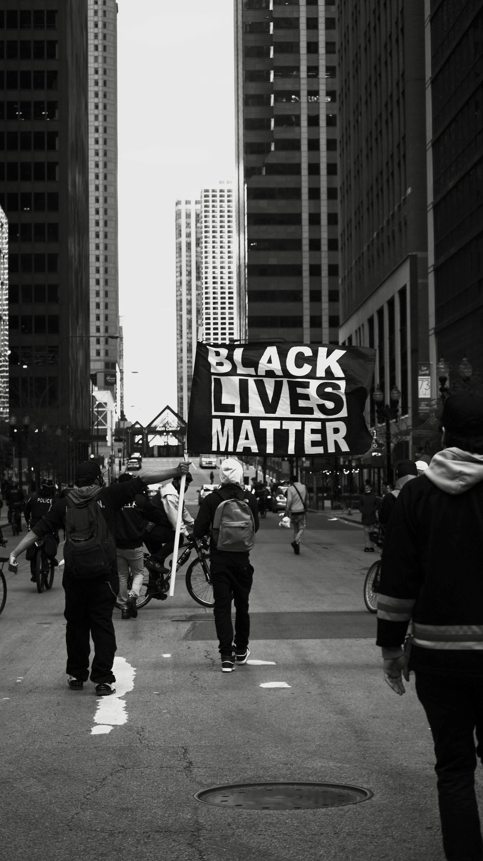 People walking and a person holding a Black Lives Matter sign in a city street with tall buildings.