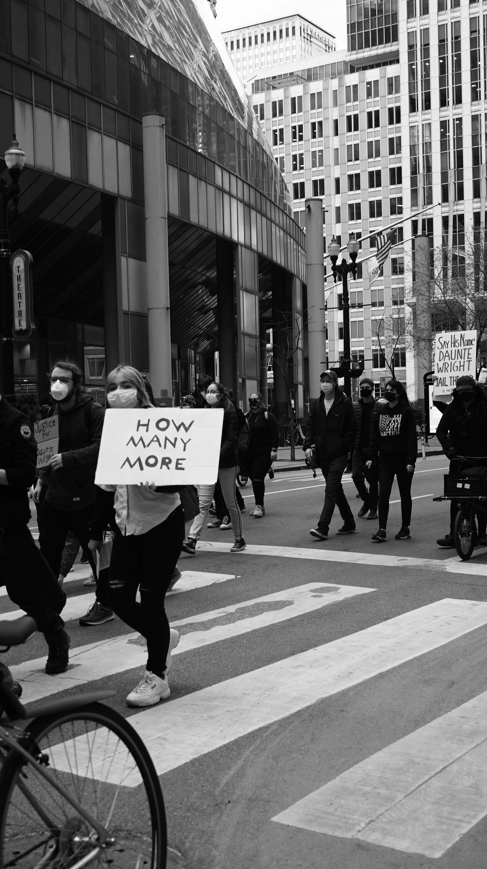 Black and white photo of a group of people walking in a city street protest, some holding signs, including one that reads 'HOW MANY MORE'; Tall modern buildings surround the scene.