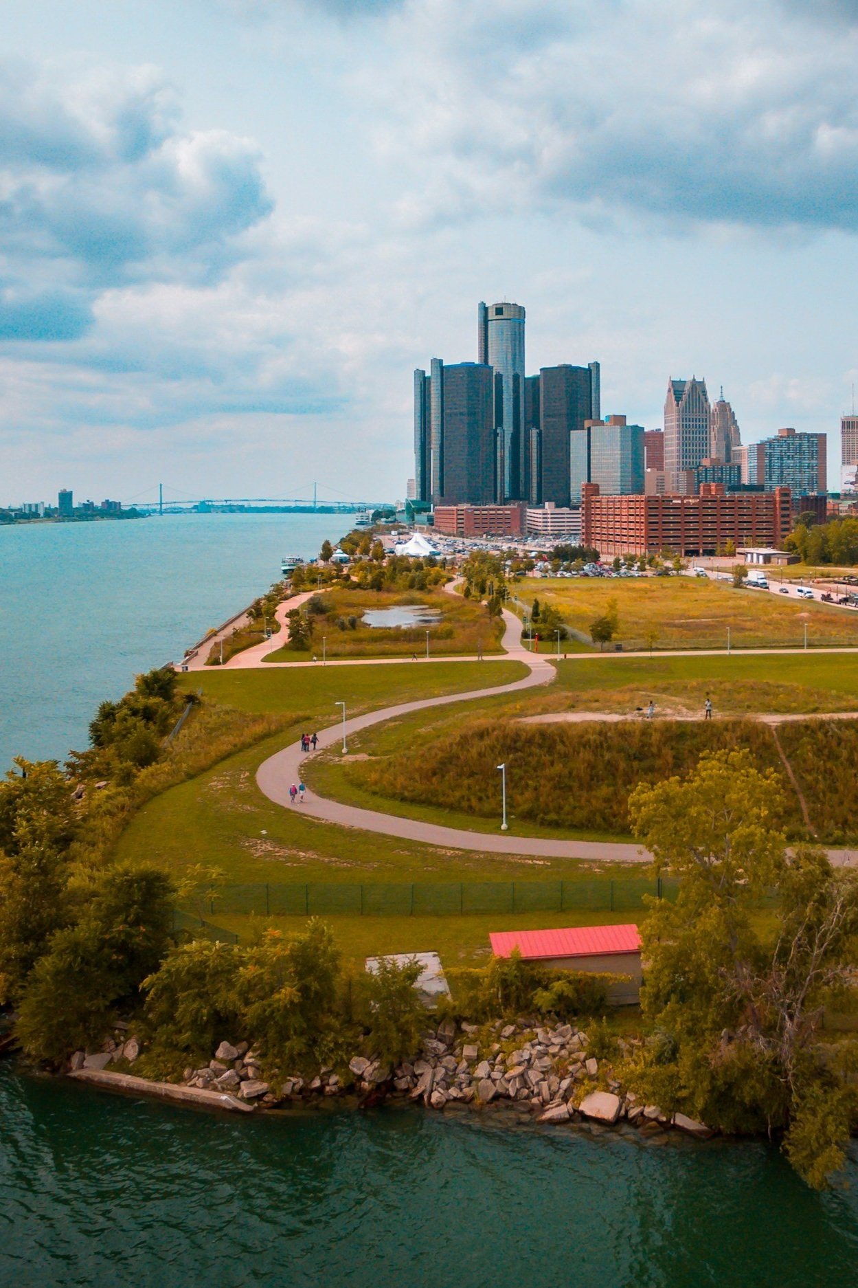 City skyline with tall buildings seen across a river, with a park in the foreground featuring winding paths, grass, trees, and a small red-roofed shelter.