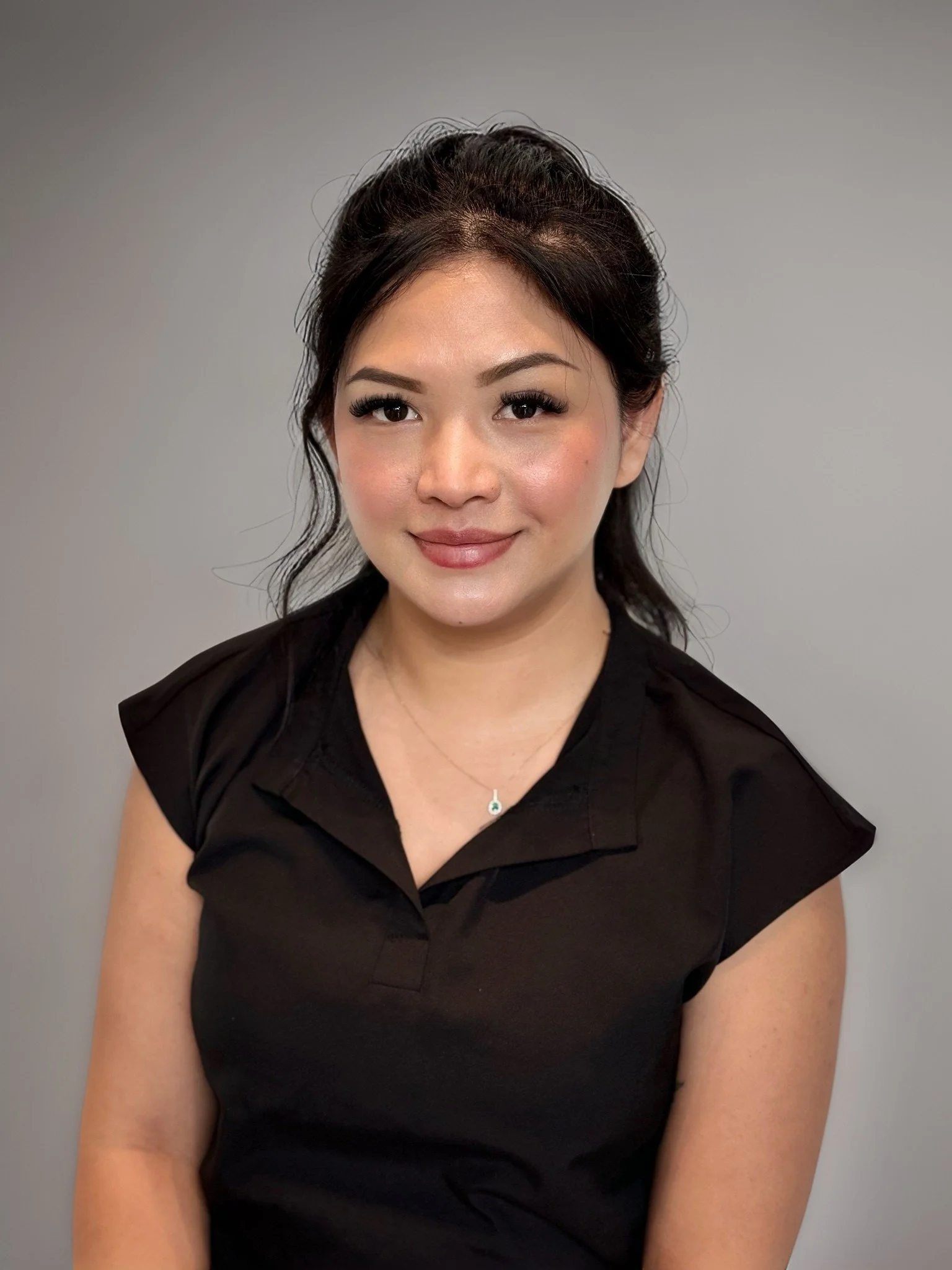 A woman with dark hair styled in an updo, wearing makeup, a black short-sleeved top, and a necklace, posing against a plain gray background.