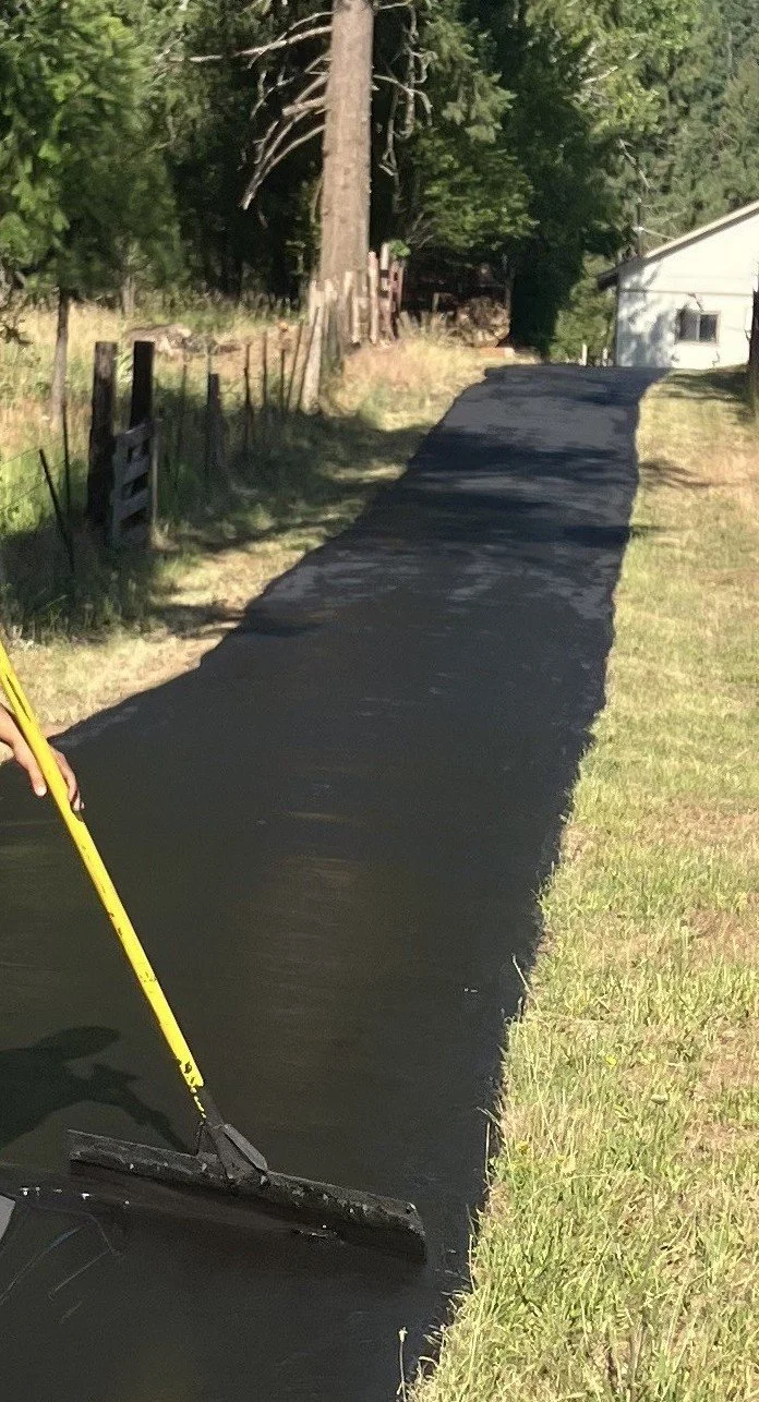 Person smoothing freshly laid black asphalt on a driveway with a heavy-duty squeegee, next to a grassy yard and a white house, with trees in the background.
