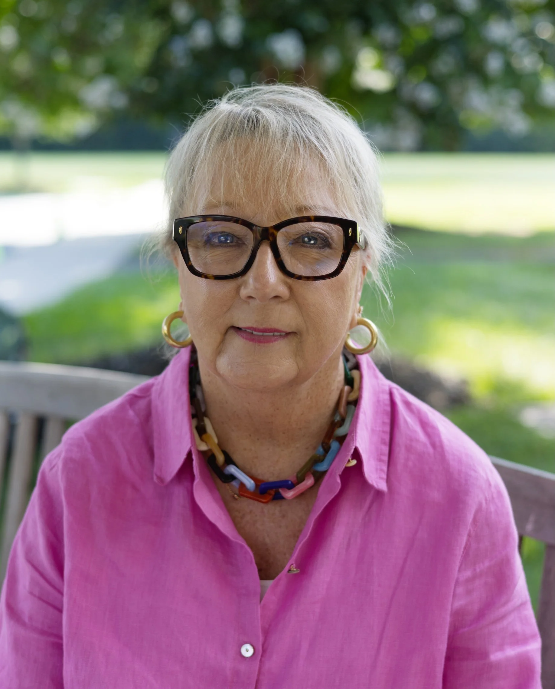 A portrait of an older woman with short blonde hair, wearing glasses, pearl earrings, a necklace, and a navy blue shirt, standing outdoors with trees in the background.