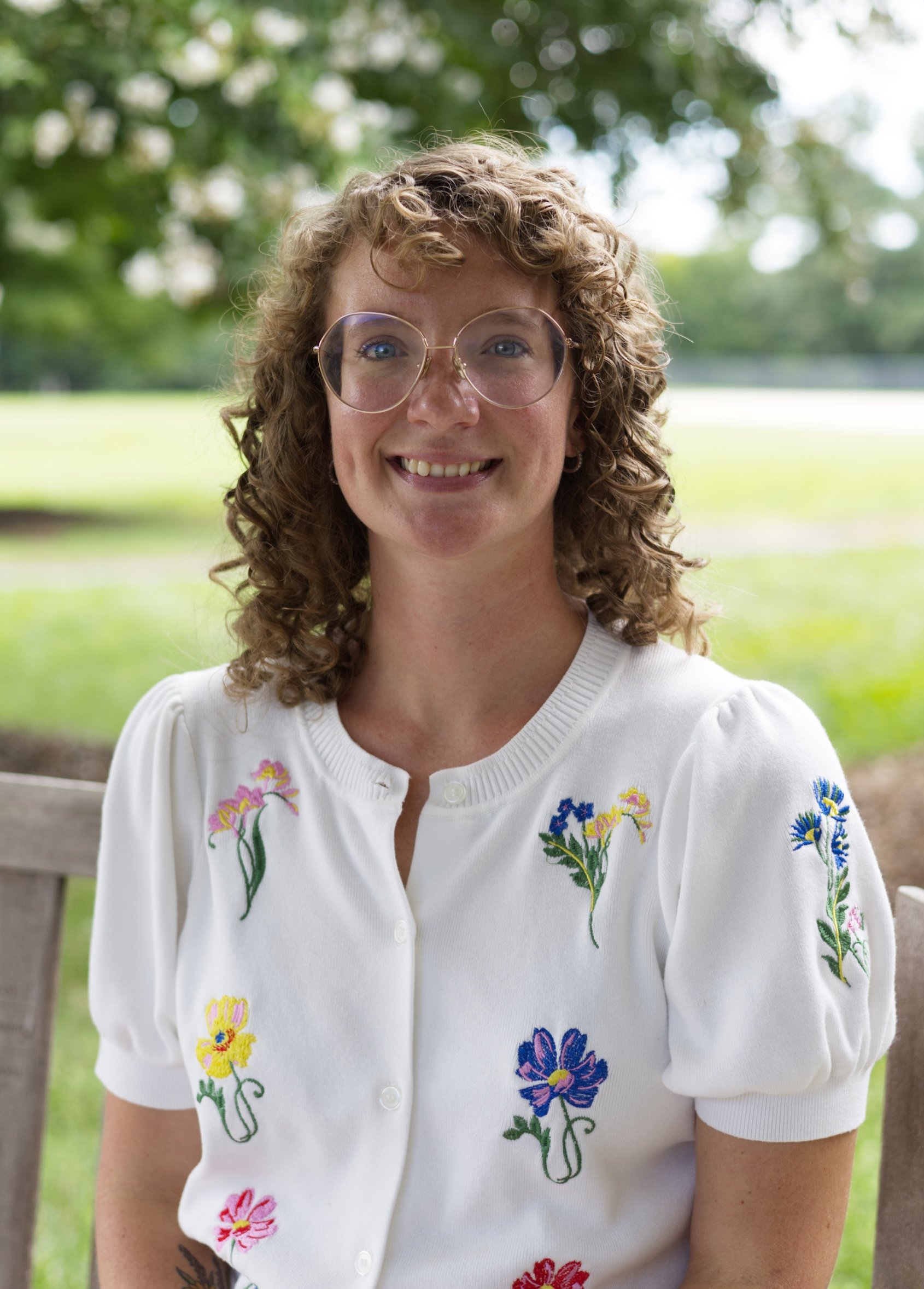 A woman with short curly brown hair and glasses, smiling outdoors with a background of trees.