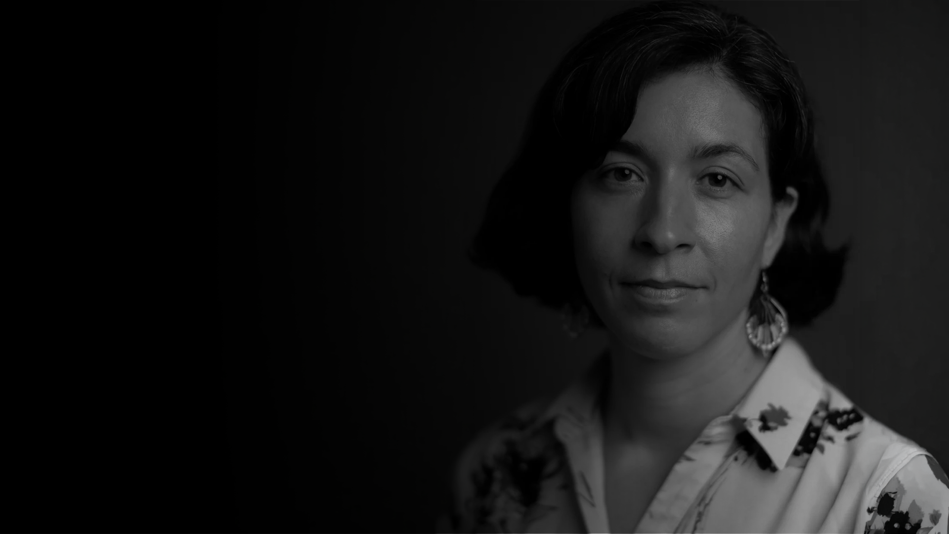 Black and white portrait of a woman with short wavy hair, wearing a floral blouse and large earrings, facing the camera with a gentle smile.