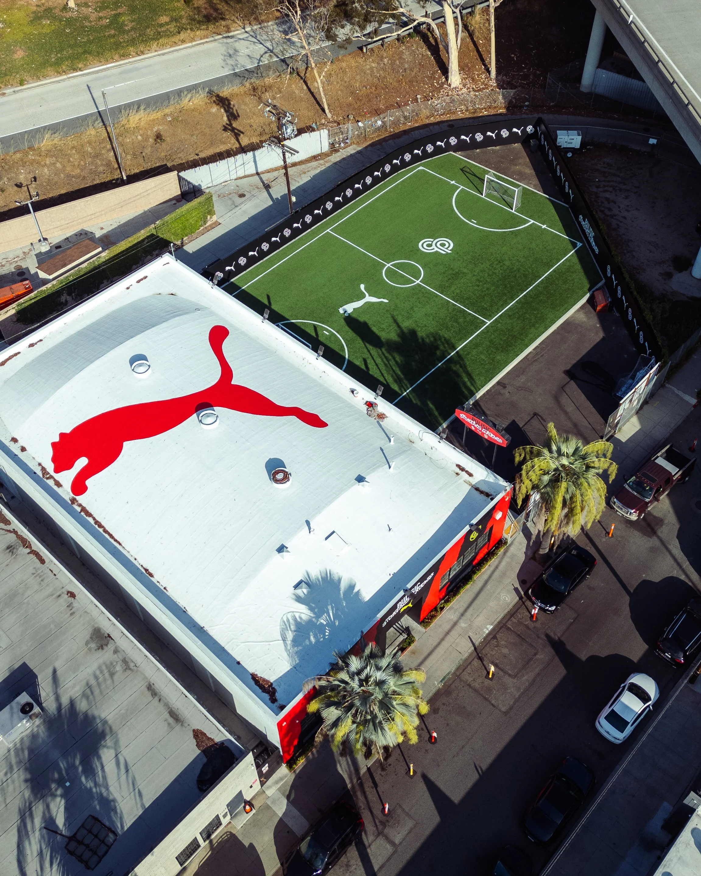 An aerial view showing two adjacent sports facilities: a rooftop with the red Puma logo and a small soccer field with artificial turf, surrounded by a black barrier with Puma logos.