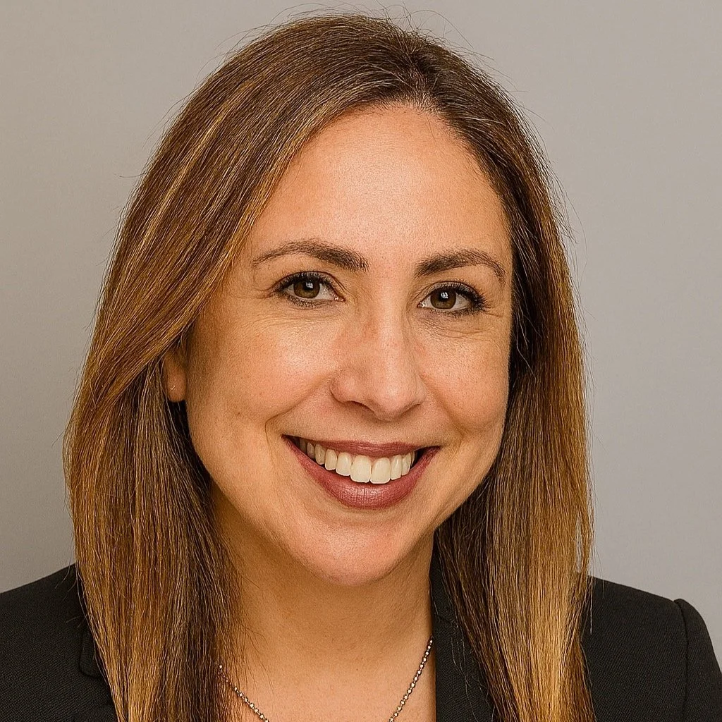 A woman with light brown hair and a broad smile, wearing a dark blazer and a necklace, against a neutral background.