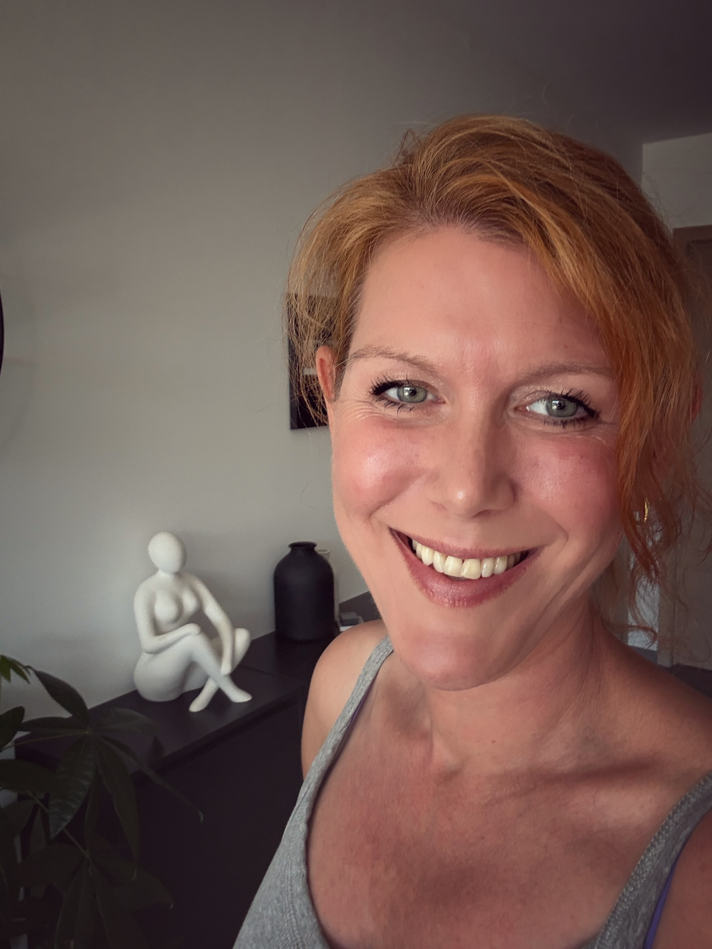 Close-up van een lachende copywriter met rood haar in een woonkamer met decoratiestukken op een kast, waaronder een witte sculptuur en een zwarte vaas, en een groene plant.