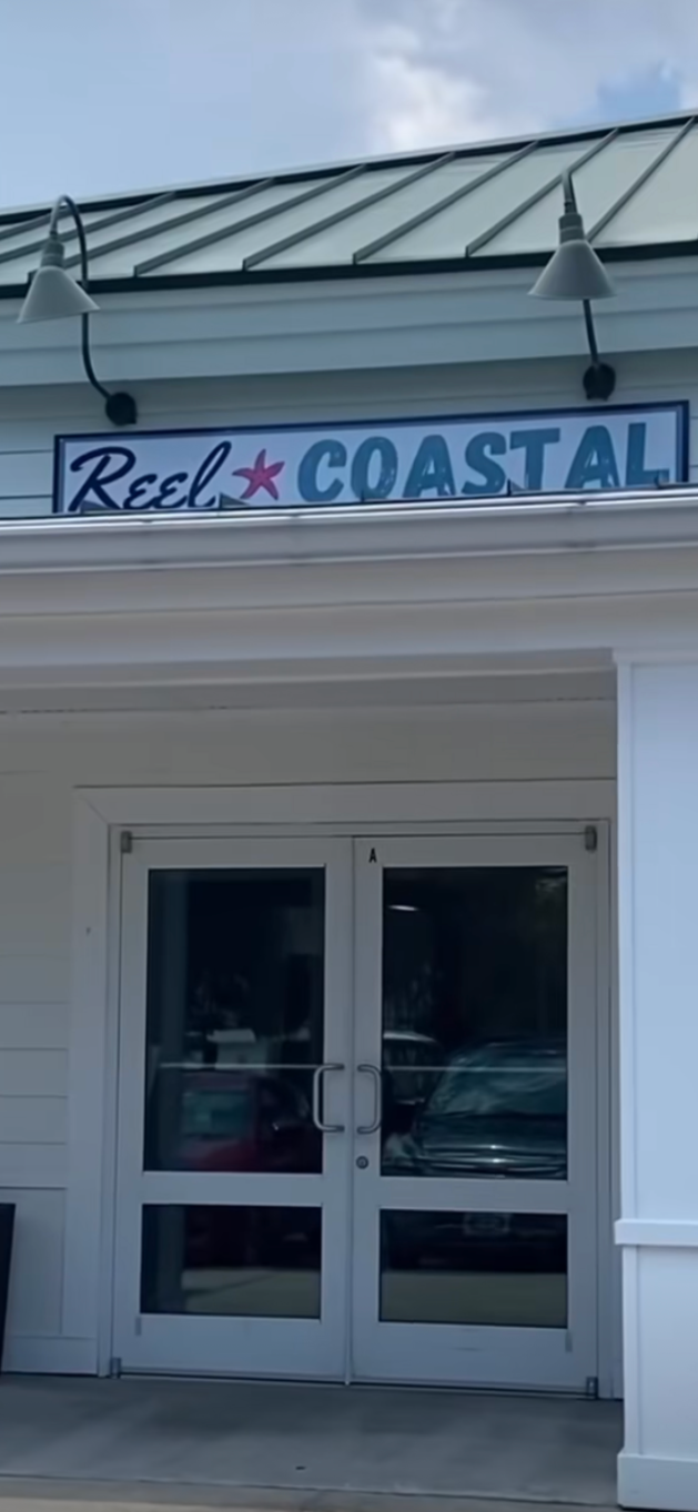 Exterior of a building with a sign that reads 'Reel Coastal', double glass front doors, and outdoor lights on the roof.