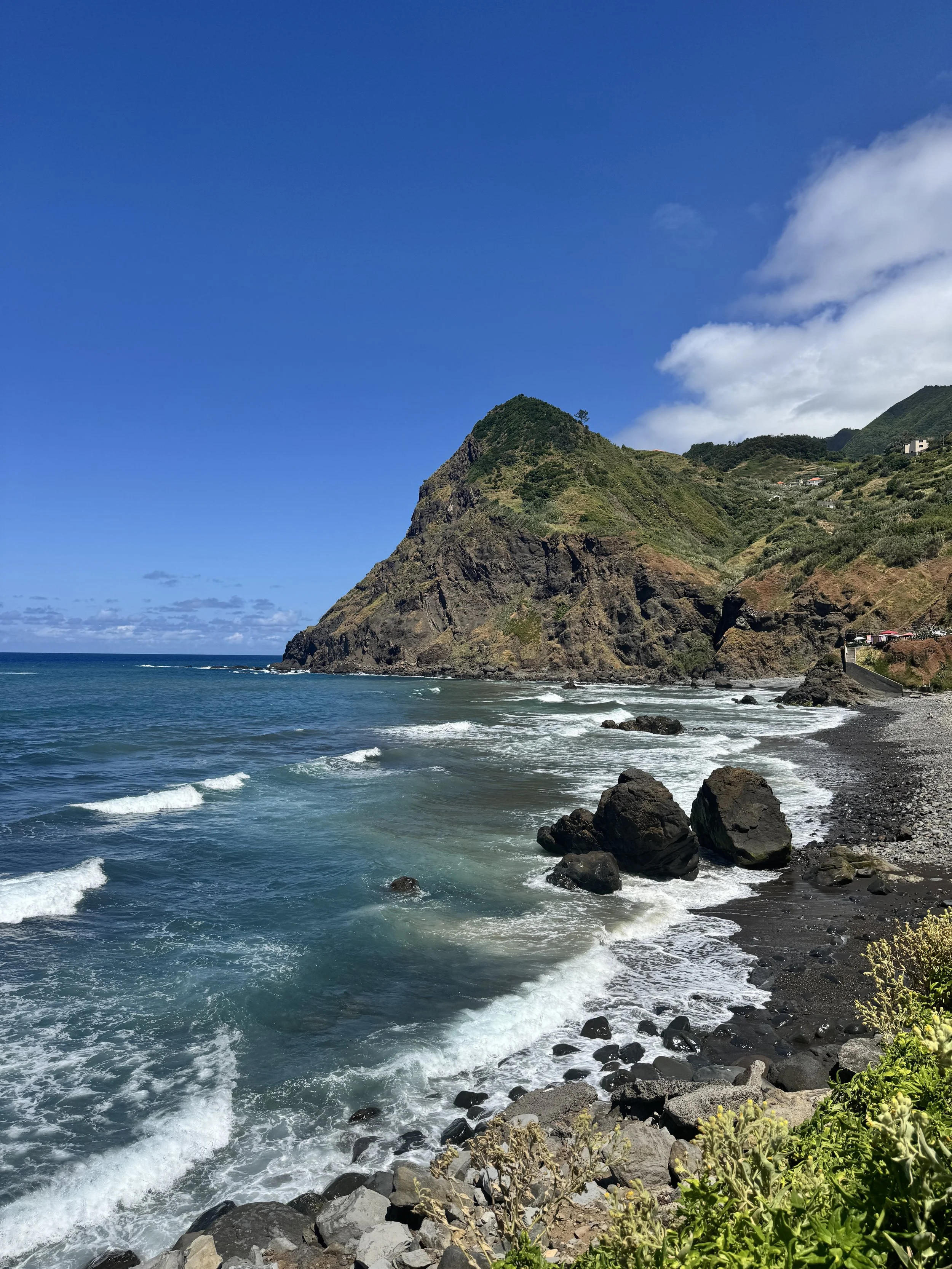 A calm gentle surf spot along Madeira’s black sand beach, where dark volcanic sand meets softly breaking Atlantic waves. The surf rolls in with an easy rhythm, creating a peaceful, understated scene against the rugged coastline of Madeira.