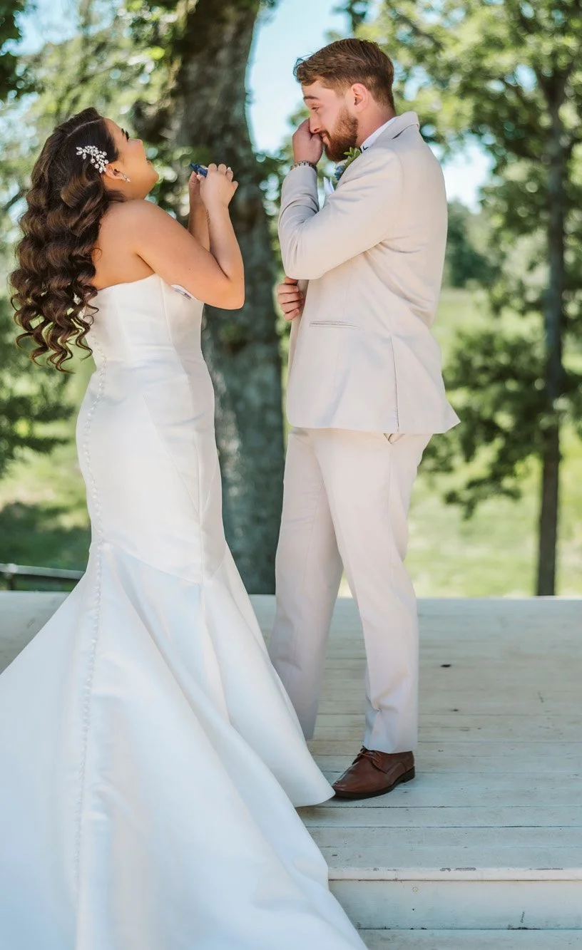 Bride and groom stand outdoors on a wooden platform with trees in the background, sharing a heartfelt moment on their wedding day.