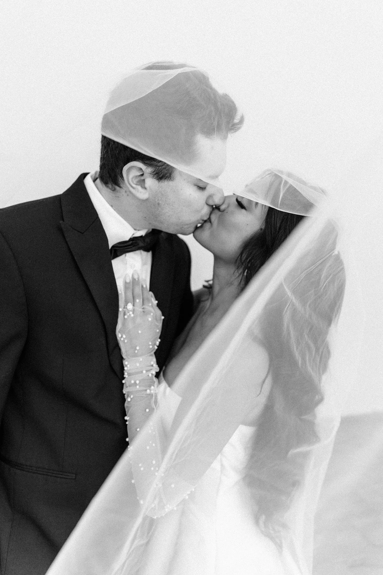 A black and white photograph of a couple kissing during their wedding. The groom is dressed in a tuxedo with a bow tie, and the bride is wearing a wedding dress with a veil and sheer gloves decorated with pearls.