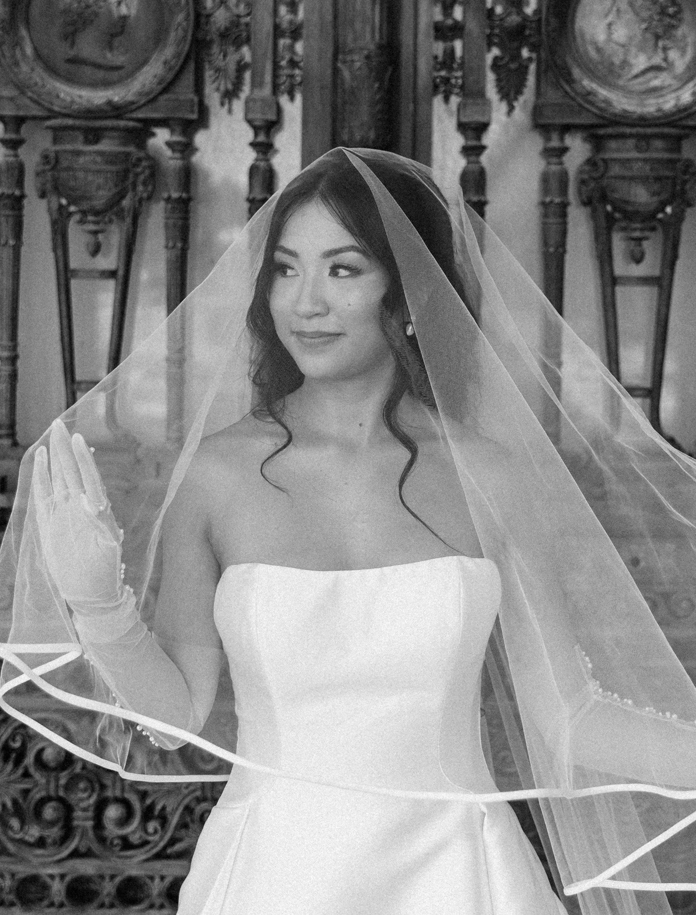 A woman in a wedding dress wearing a veil, standing in an ornately decorated room, looking to the side.