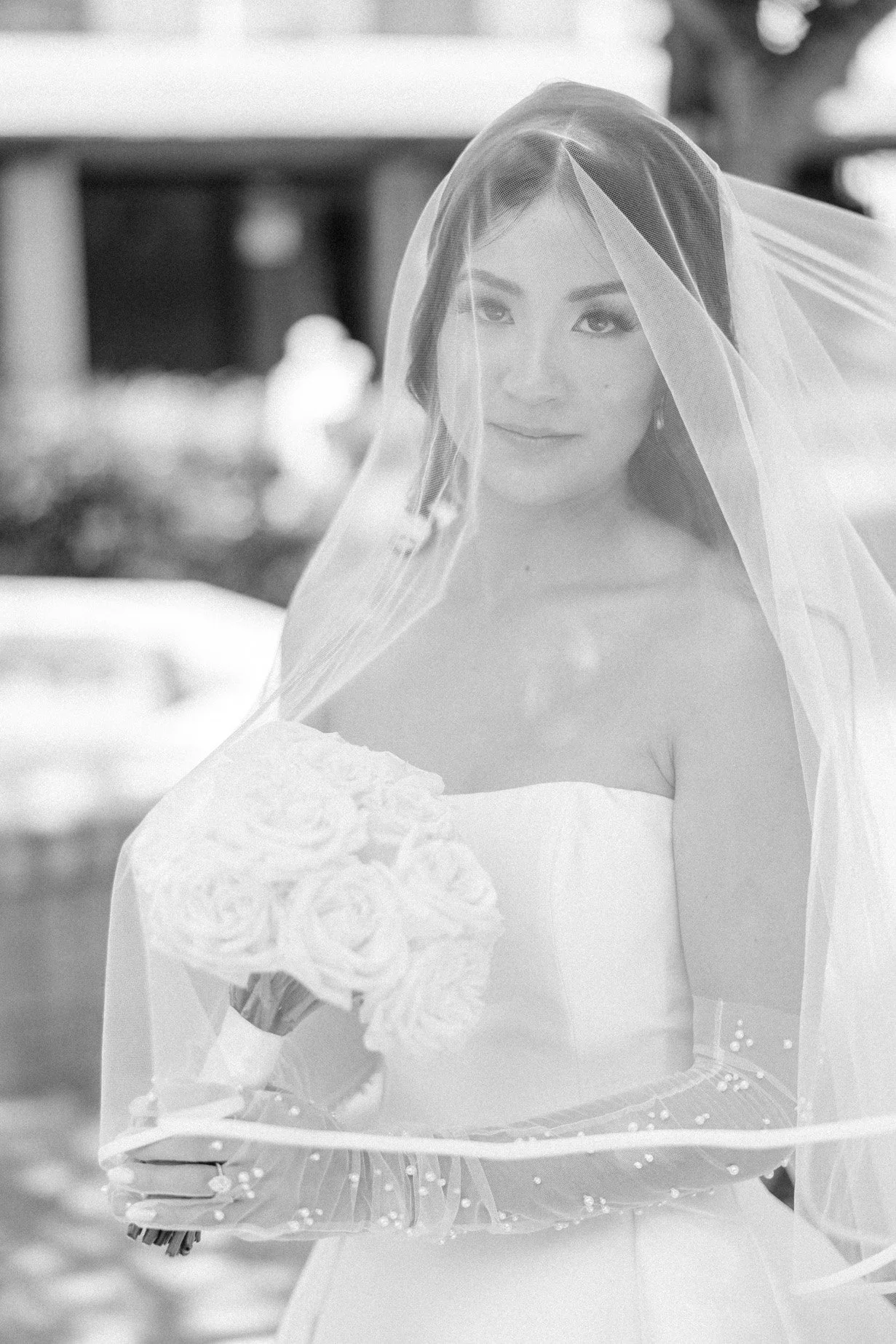 A bride with dark hair and a veil holding a bouquet of roses, wearing a strapless wedding dress, standing outdoors.