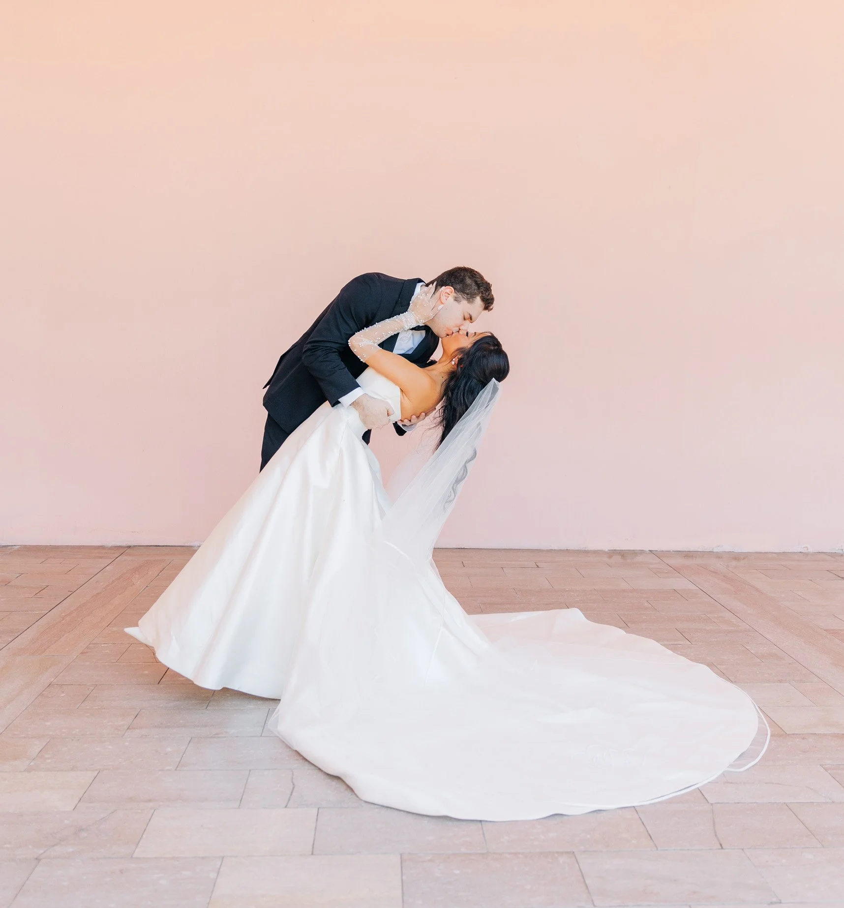 A bride and groom dancing together. The groom is dipping the bride, who is wearing a long white wedding gown with a veil, while the groom is in a black tuxedo. The background is a plain light pink wall.