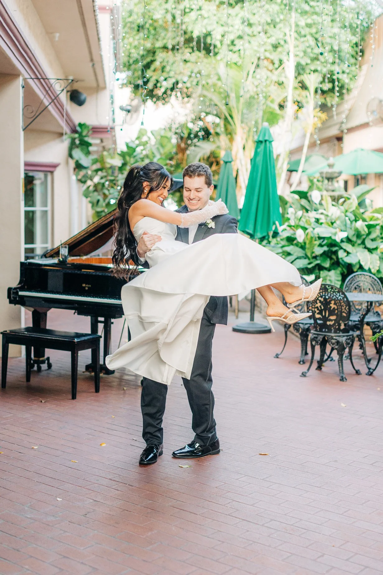 A groom in a black tuxedo lifting a bride in a white dress and heels in an outdoor setting, with greenery and patio furniture in the background.