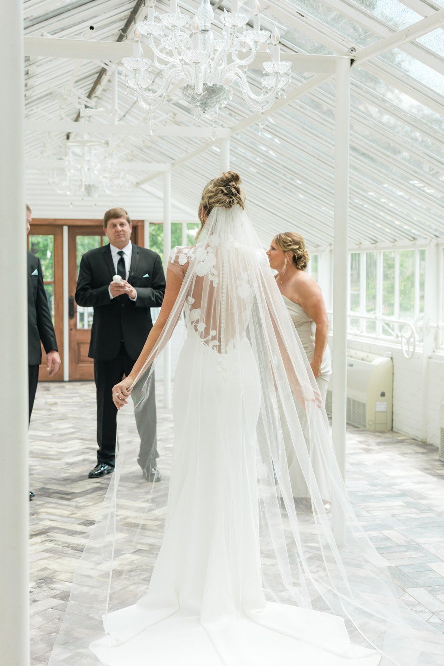 Bride in a wedding dress with a veil, standing inside a bright, glass-enclosed venue, surrounded by people in formal attire.
