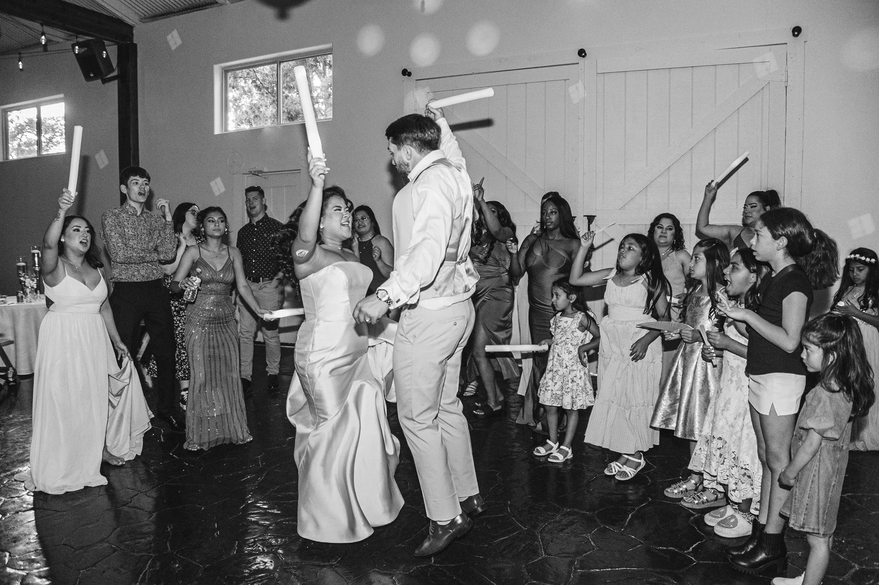 A black and white photo of a lively celebration with many children and adults dancing and holding glow sticks or foam sticks in a decorated room with barn doors and windows.