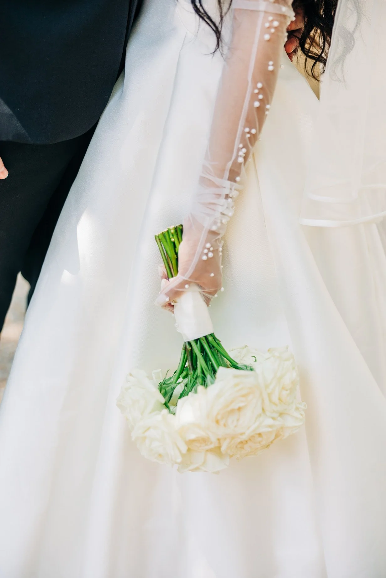 A bride in a white wedding dress holding a bouquet of white roses with a man in a suit beside her, possibly during a wedding ceremony.