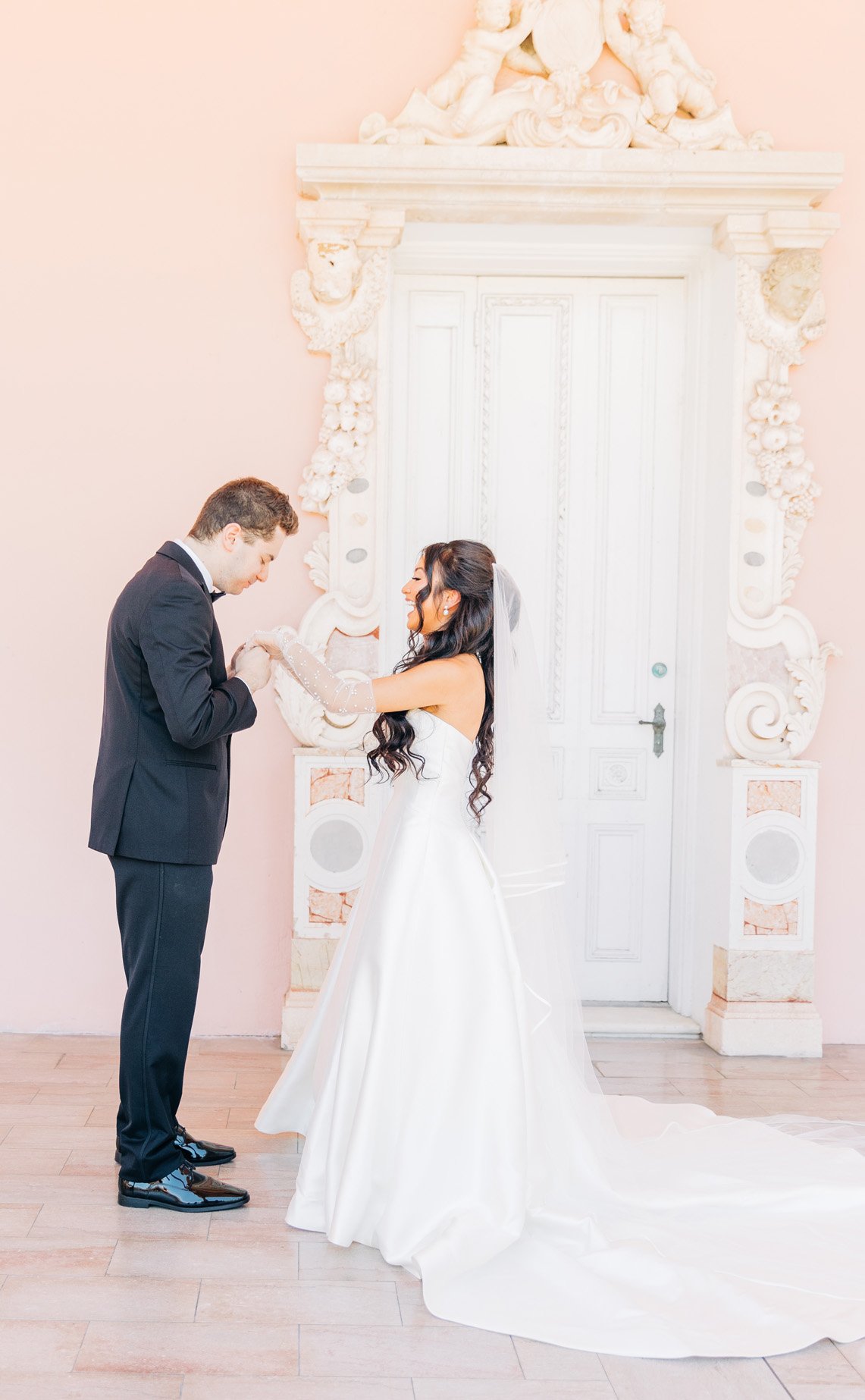 A bride and groom exchanging vows in a room with pink walls and ornate white decor.