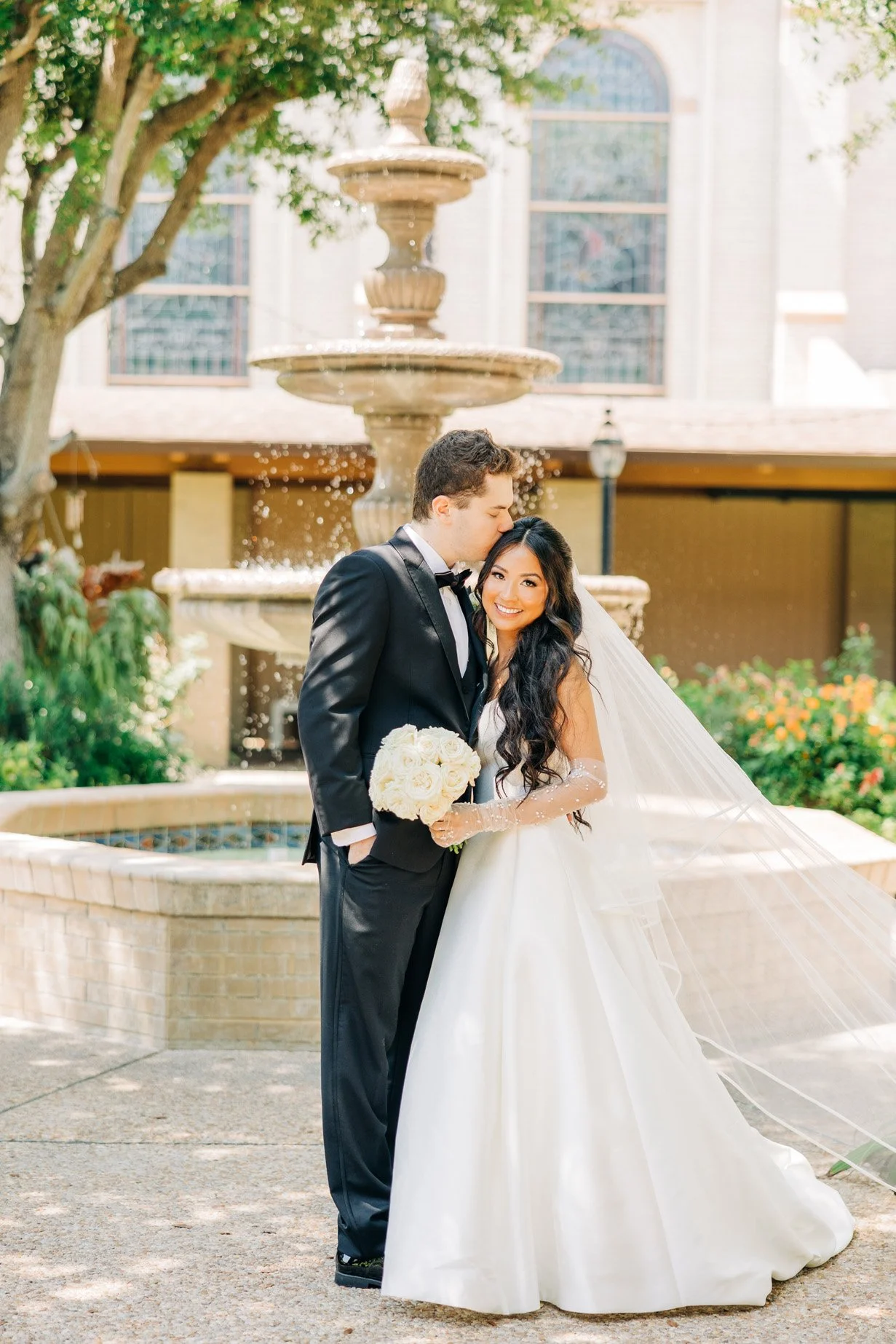 Bride and groom posing near a fountain with the groom kissing the bride on the forehead, outdoor wedding photoshoot.