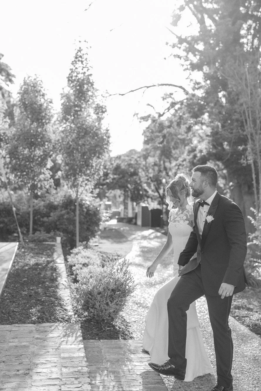 A black and white photo of a newlywed couple walking outdoors on a sunny day, surrounded by trees and bushes.