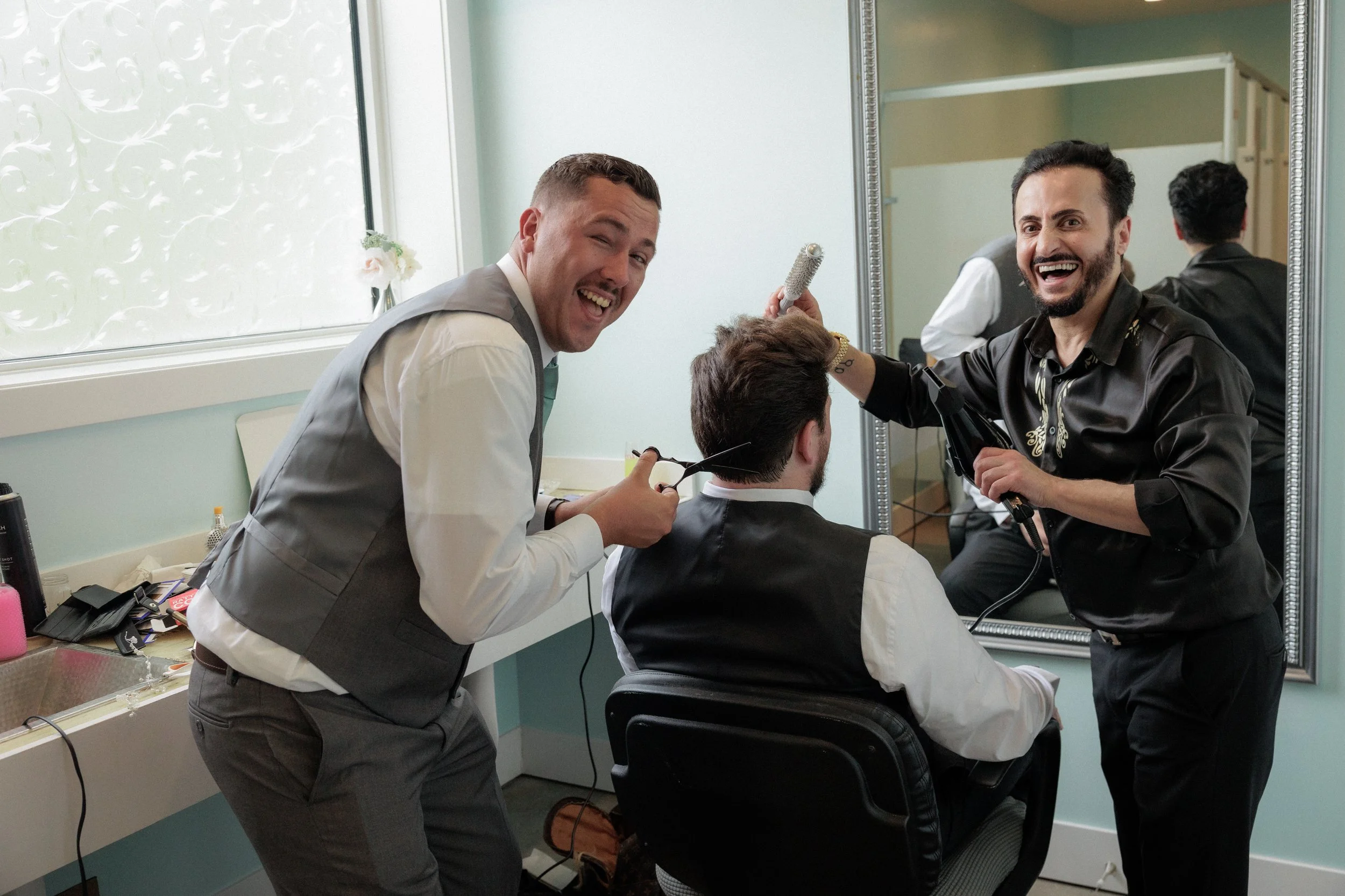 Two men in suits hairstyling a seated man with dark hair, using scissors and a hairbrush, in a mirror reflection in a bright, modern salon.