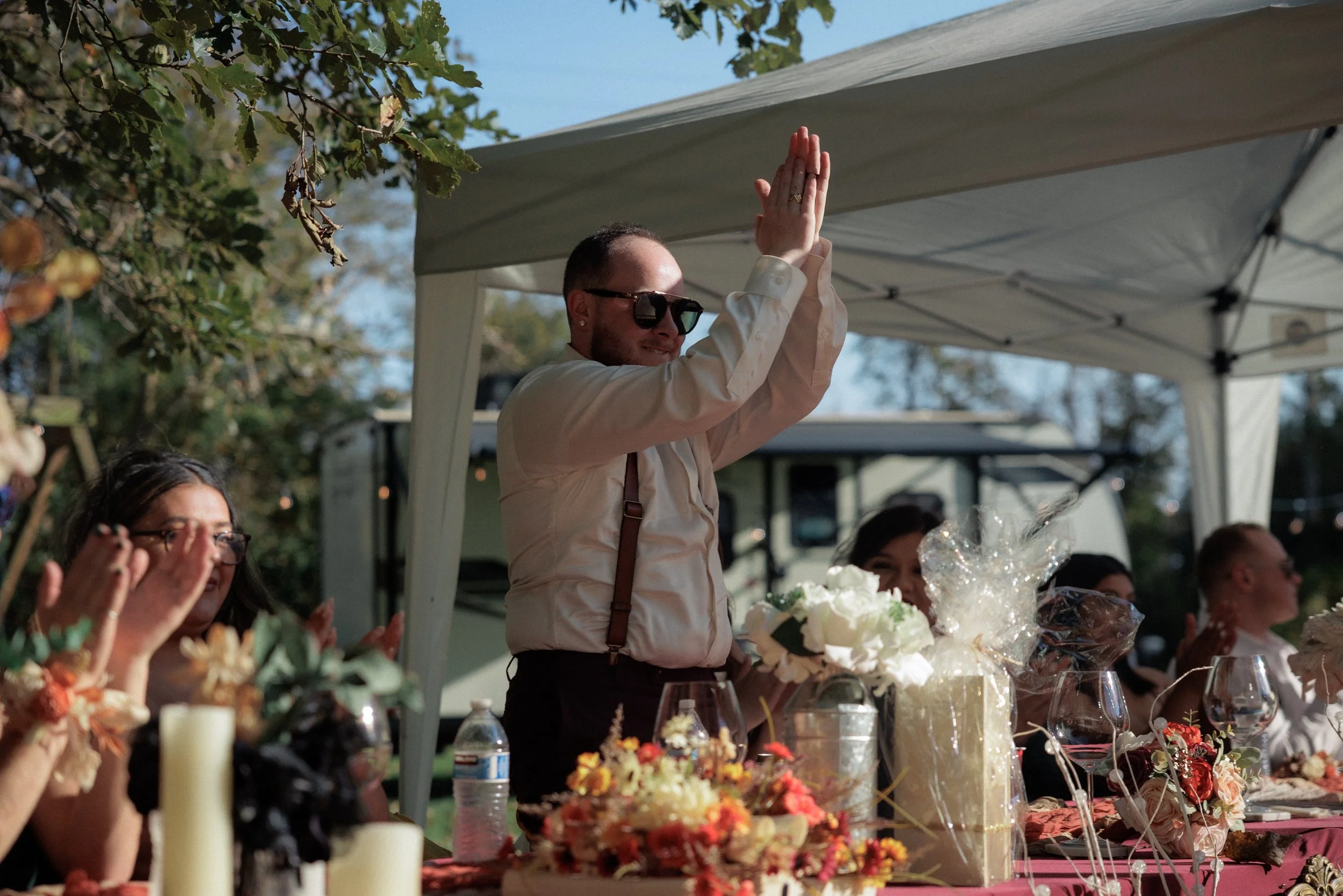 A man in a beige shirt and sunglasses is clapping at an outdoor celebration under a white canopy, with a table of flowers, gifts, and candles in front of him.