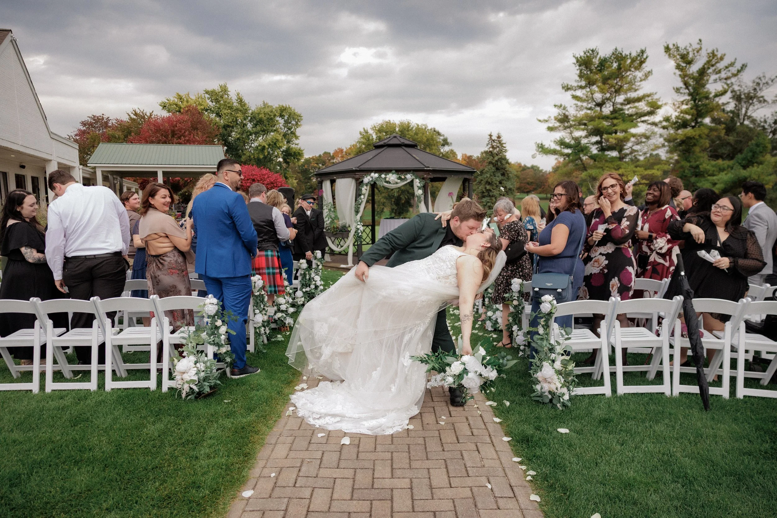 A newlywed couple sharing a kiss outdoors at their wedding ceremony, with guests standing and watching on either side of the aisle decorated with white flowers and greenery, under a cloudy sky.