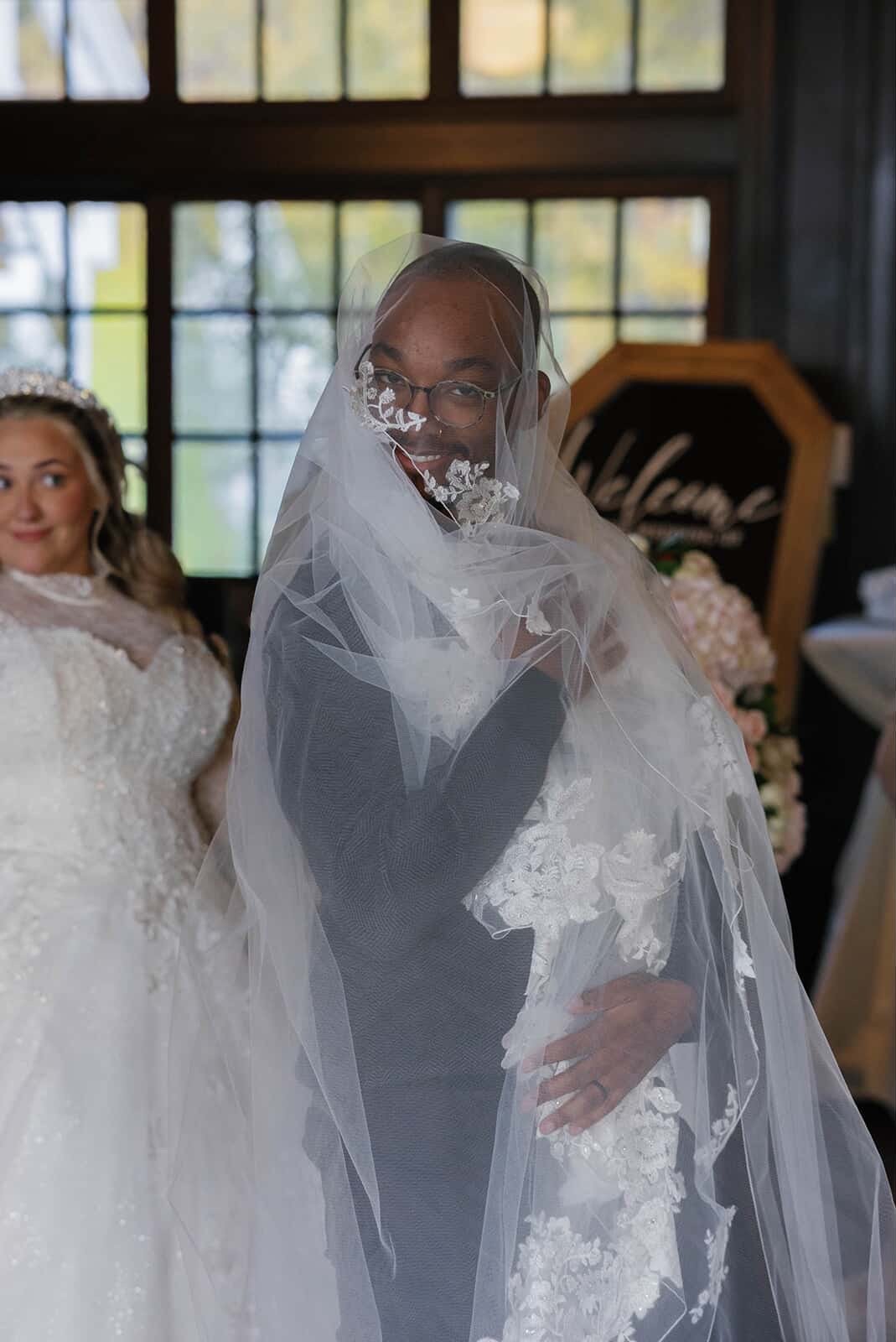 A bride wearing glasses, a black dress, and a wedding veil, smiling at the camera, standing in a venue with large windows. A maid of honor or bridesmaid in a white dress with lace details is visible in the background.