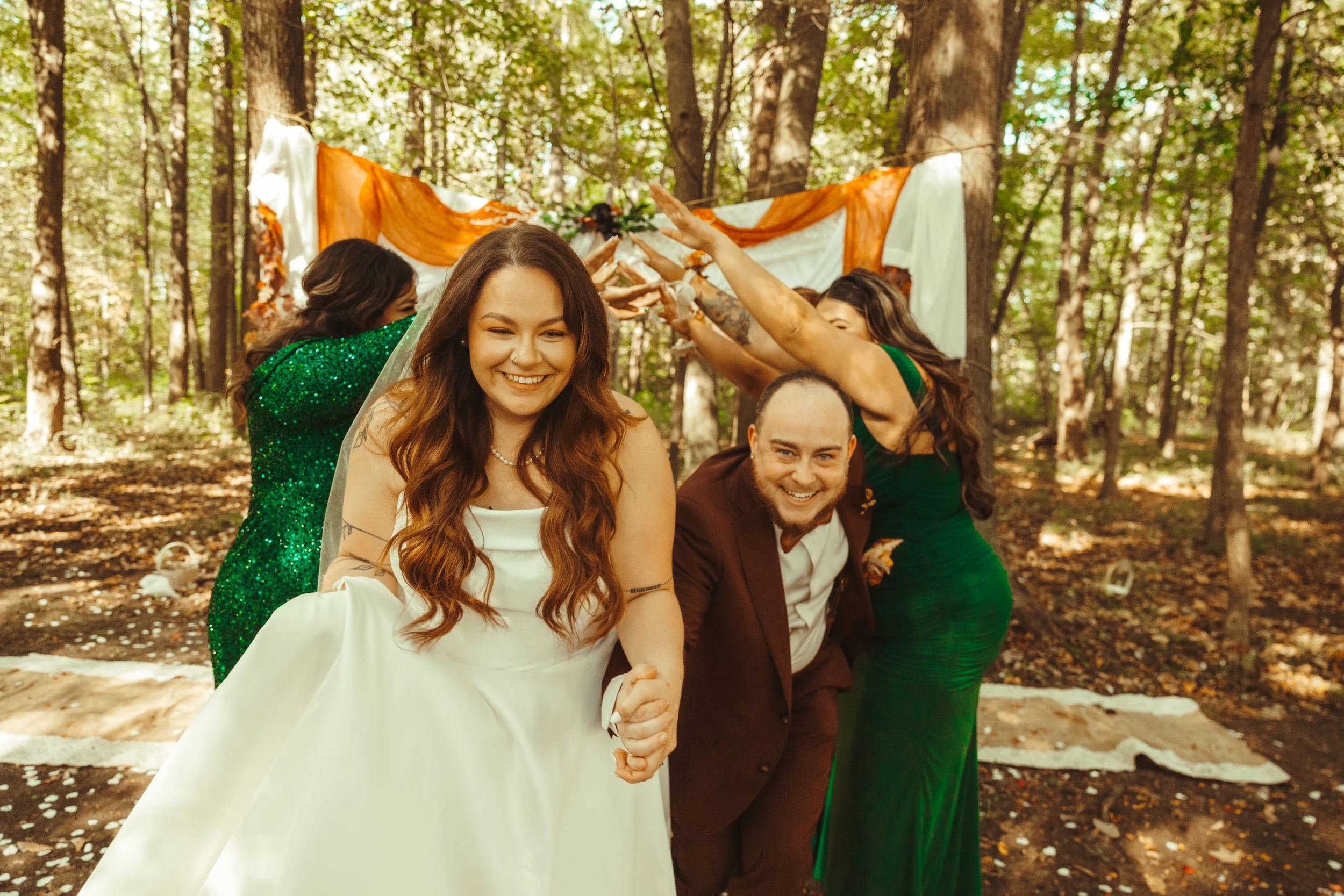 A wedding ceremony outdoors in a forest with a bride, groom, and two women. The bride is smiling, holding hands with the groom, and wearing a white wedding dress. The groom is in a burgundy suit, crouching, and smiling. Two women in green dresses are participating in a handfasting ritual with a cloth, surrounded by trees and autumn leaves.