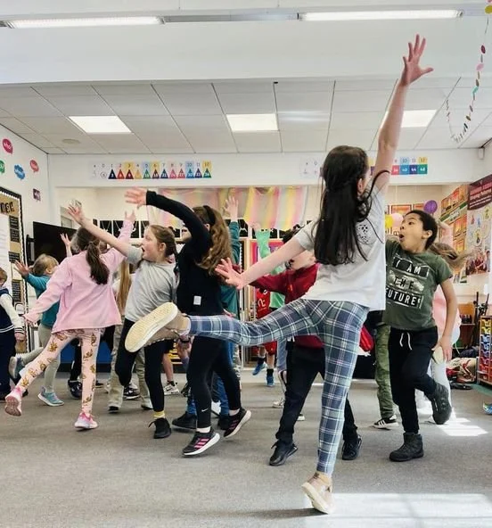 Group of children dancing energetically in a classroom with colorful decorations and posters on the walls.