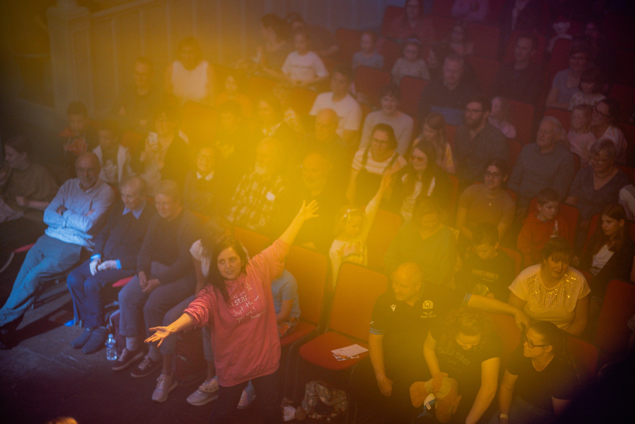 Audience at a theater performance with a woman in pink interacting with the crowd.