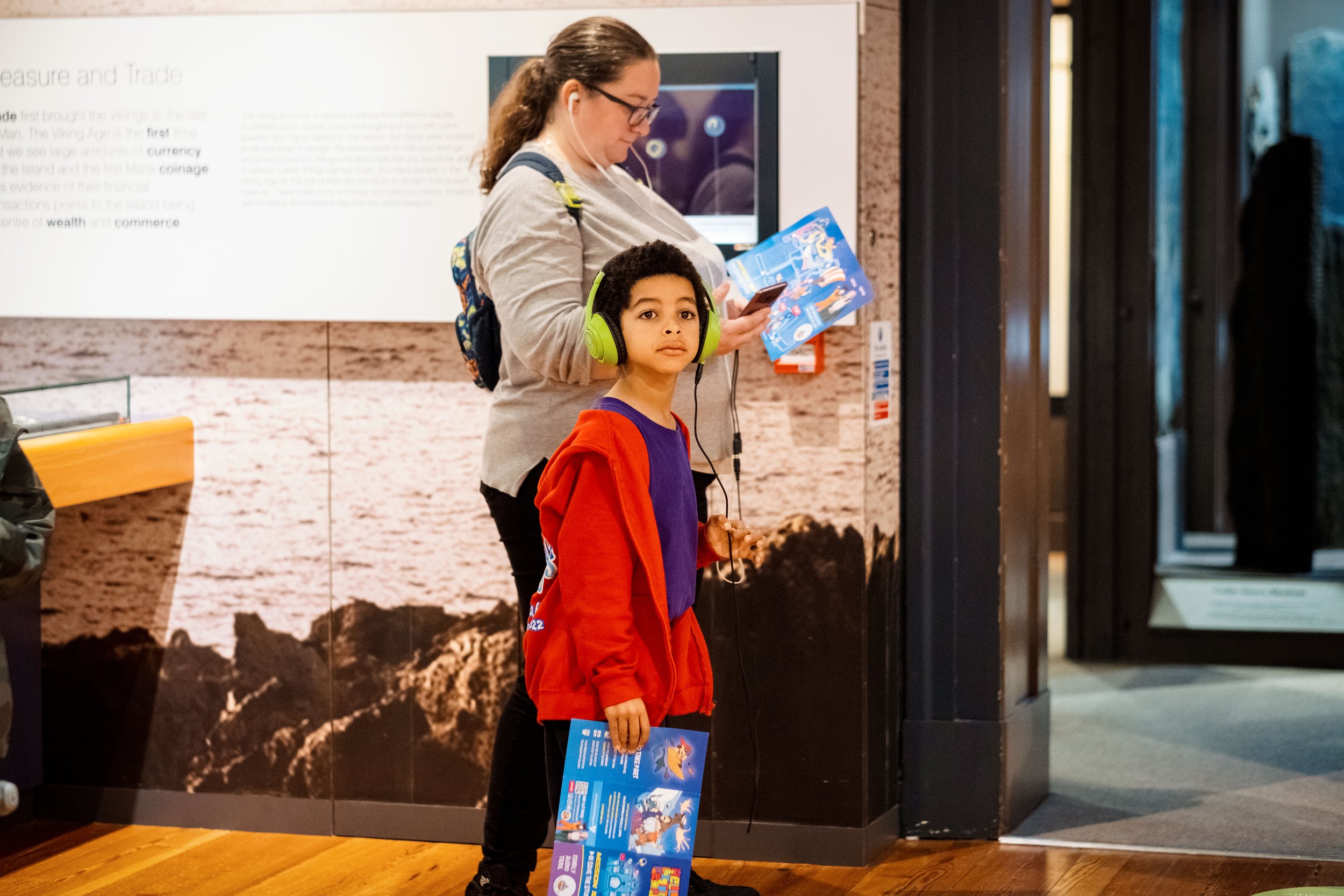 Child wearing green headphones holding a booklet in a museum setting with an adult looking at a phone.