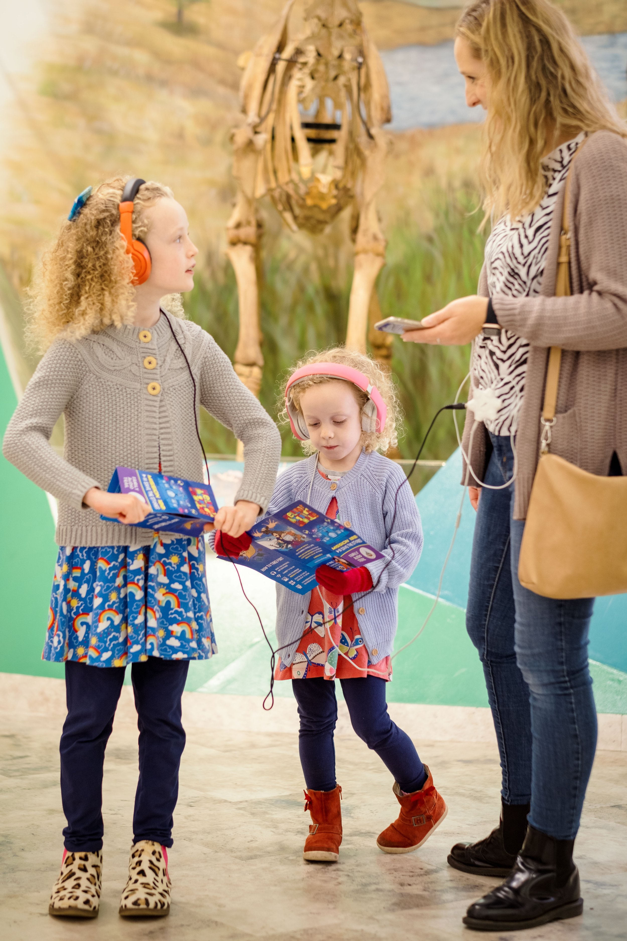 Two young children wearing headphones are looking at brochures in a museum. An adult with a smartphone stands next to them. A dinosaur skeleton is in the background.