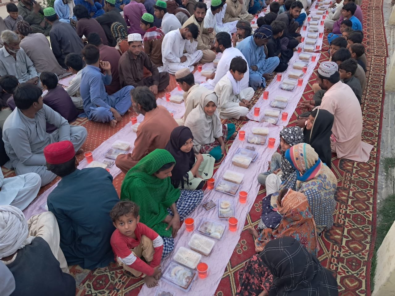 Group of people gathered for a communal meal, seated on carpets, with food trays and drinks in cups placed in front of them.