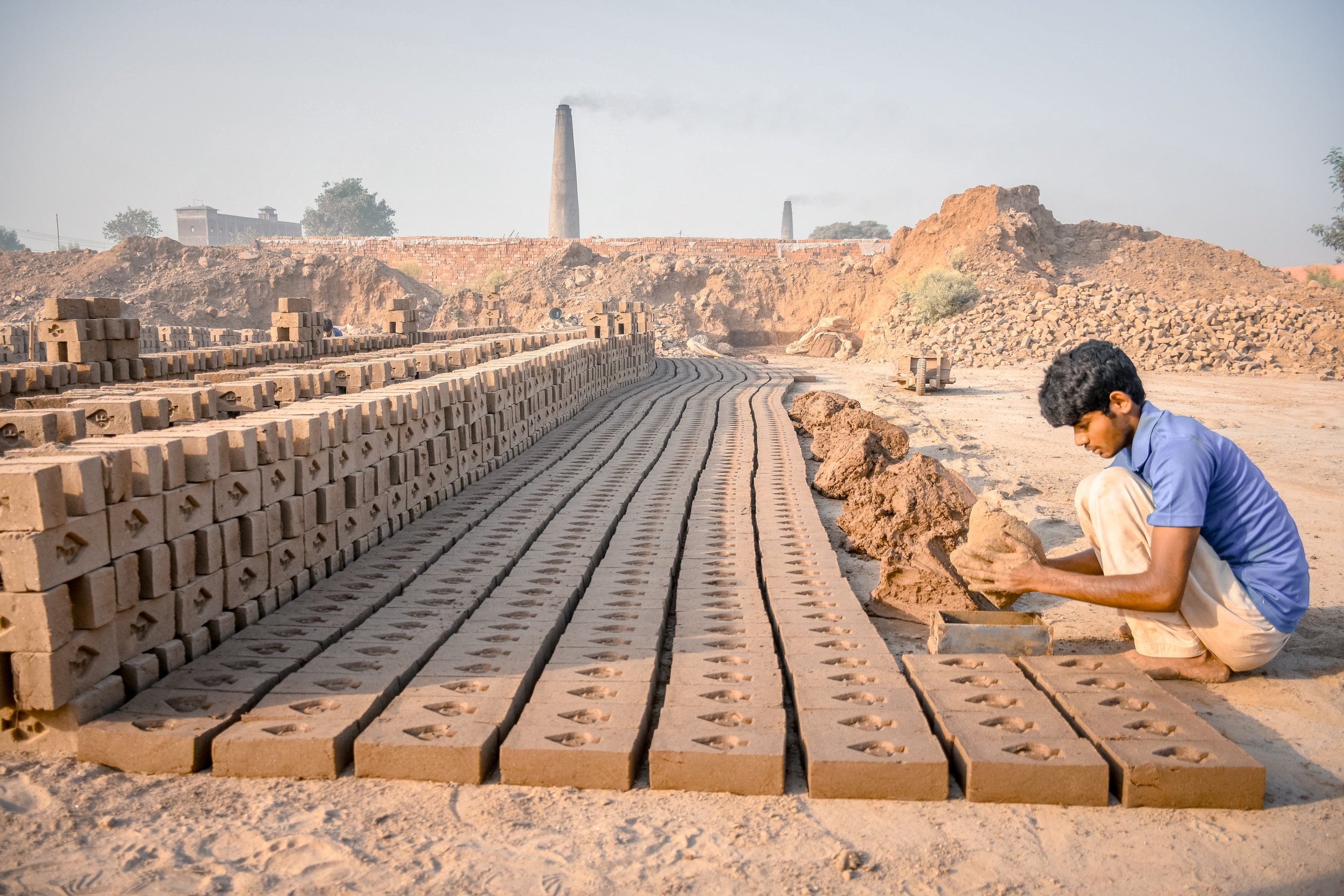 Worker making clay bricks in a brick kiln, with stacks of bricks drying outs. Smokestacks in the background.