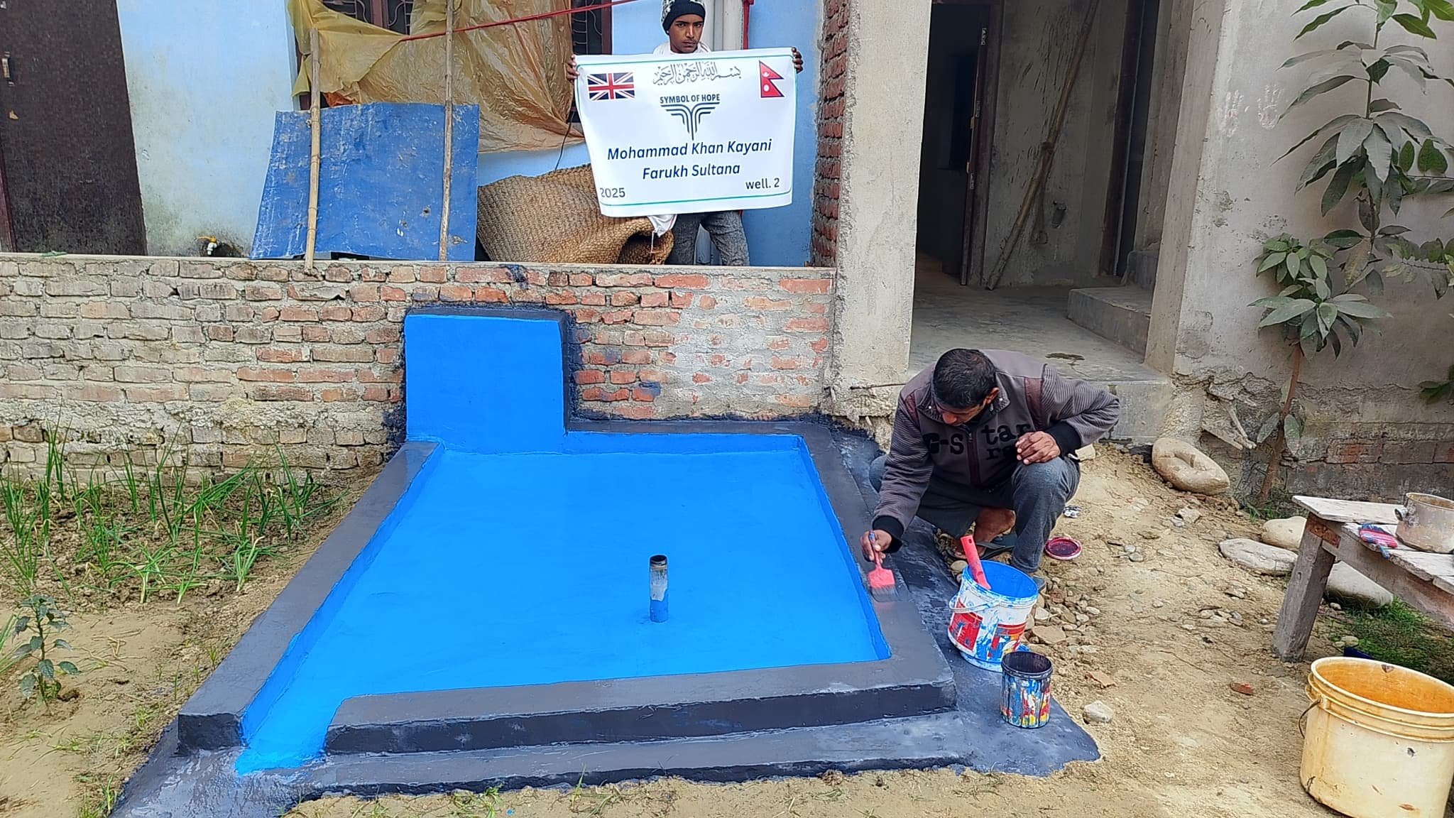 Man painting a blue rectangular structure outdoors next to a brick wall; another person holds a banner. Buckets of paint on the ground.